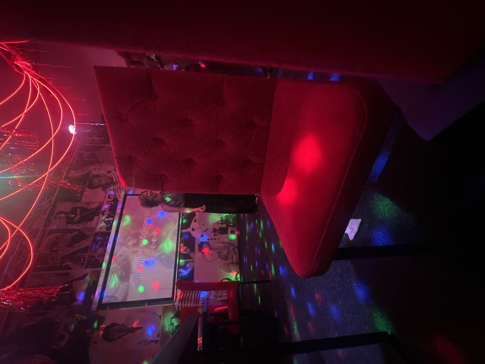The image depicts an indoor karaoke bar, likely located in Chaikovsky, Russia, given the text on the screen. The scene is dimly lit, with vibrant red neon lights and colorful disco lights casting a festive atmosphere. In the foreground, a plush red armchair dominates the frame, its tufted design and rich color suggesting a comfortable seating arrangement. Behind it, a large projection screen displays lyrics in Russian, indicating a karaoke session is in progress. A woman, her face partially visible, stands near the screen, appearing to be participating in the karaoke. The walls are adorned with posters and black and white images, adding to the retro ambiance. The floor is dark and appears to be carpeted, with scattered patches of colored light reflecting on its surface. The overall impression is one of a lively and entertaining night out.