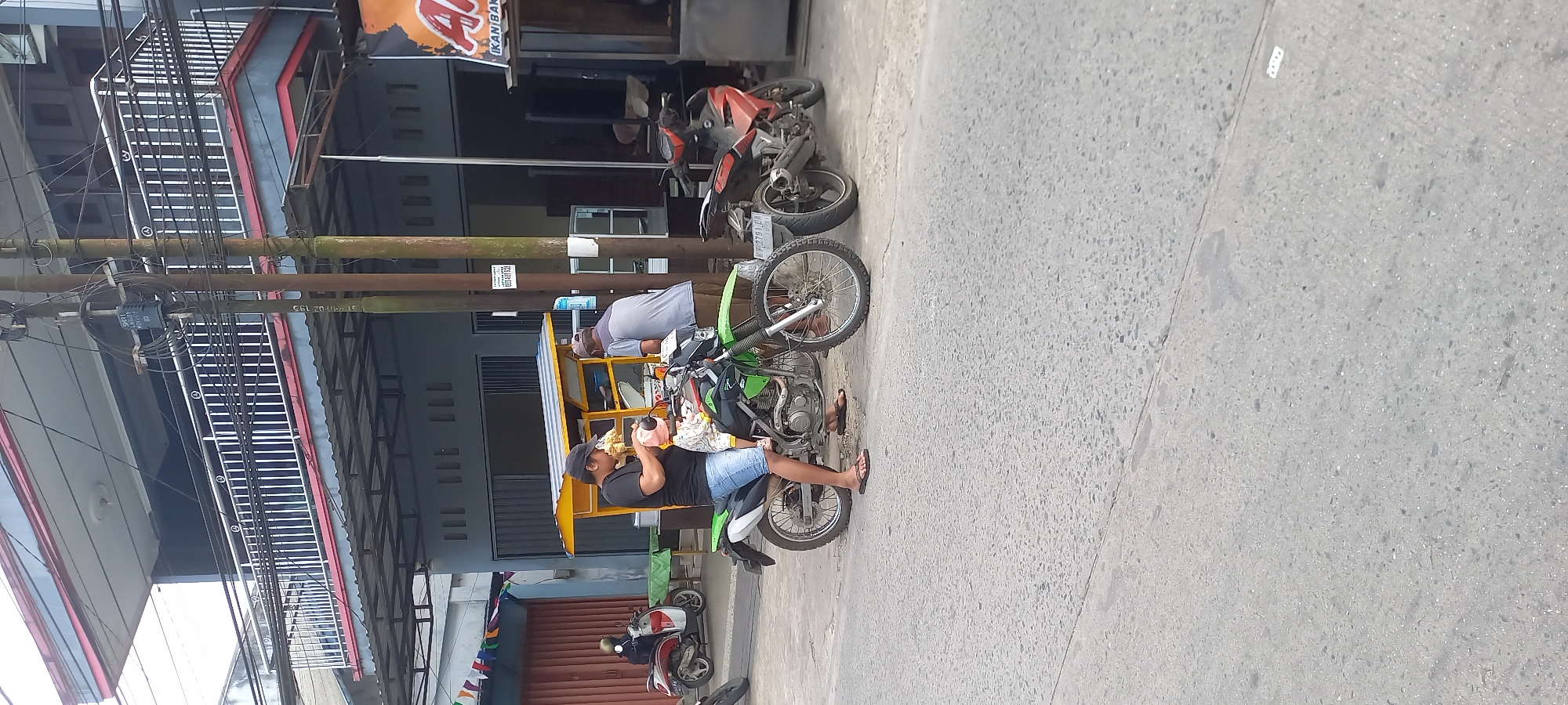 The image depicts a street scene in Samarinda, Indonesia, with a focus on a man sitting on a green dirt bike. He is wearing a black t-shirt, blue denim shorts, and flip-flops. In his hands, he holds a light-colored object, possibly a baby bottle, and appears to be feeding a baby. Next to the green dirt bike, a red motorcycle is parked. In the background, there is a building with a balcony and an outdoor food stall. The stall has a yellow and blue awning and appears to be serving food. A man in a grey t-shirt and brown pants is standing behind the stall. Several electric poles with wires are also visible, as are small triangular flags strung across the area. The ground is paved with asphalt.