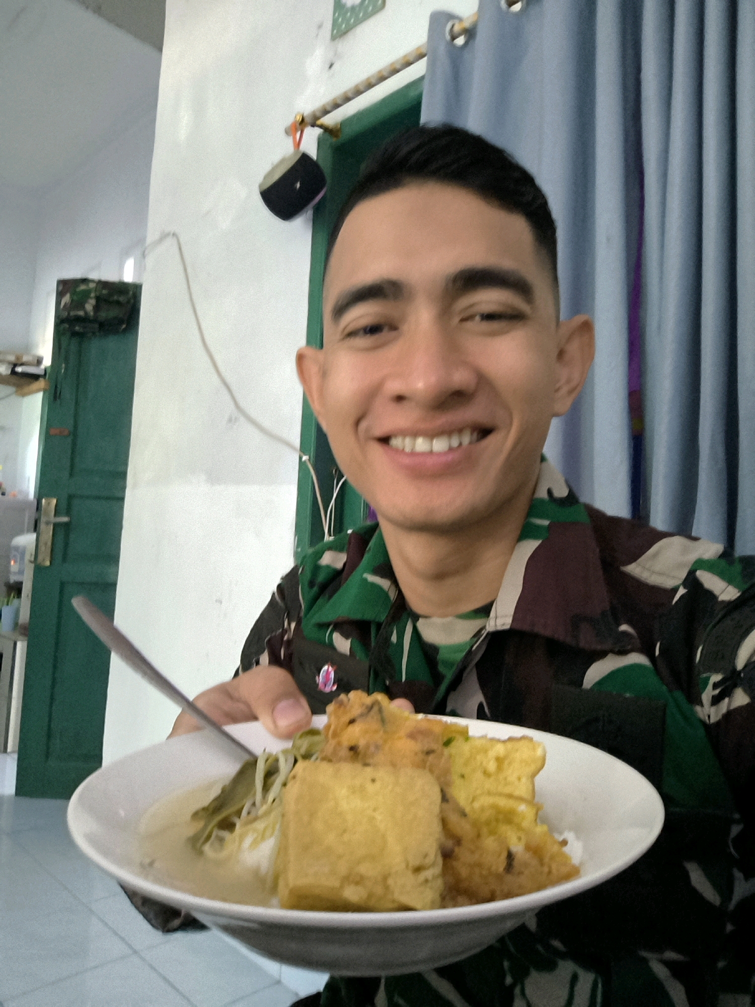 A young man in a camouflage military uniform smiles at the camera while holding a white bowl of food. The food consists of rice, what appears to be fried tofu, and some vegetables. He is holding a spoon in his right hand, about to take a bite. The background suggests an indoor setting with a white wall, a green door, and blue curtains. The overall scene is casual and appears to be a mealtime in what could be barracks or a military living quarter in Unknown, Indonesia.