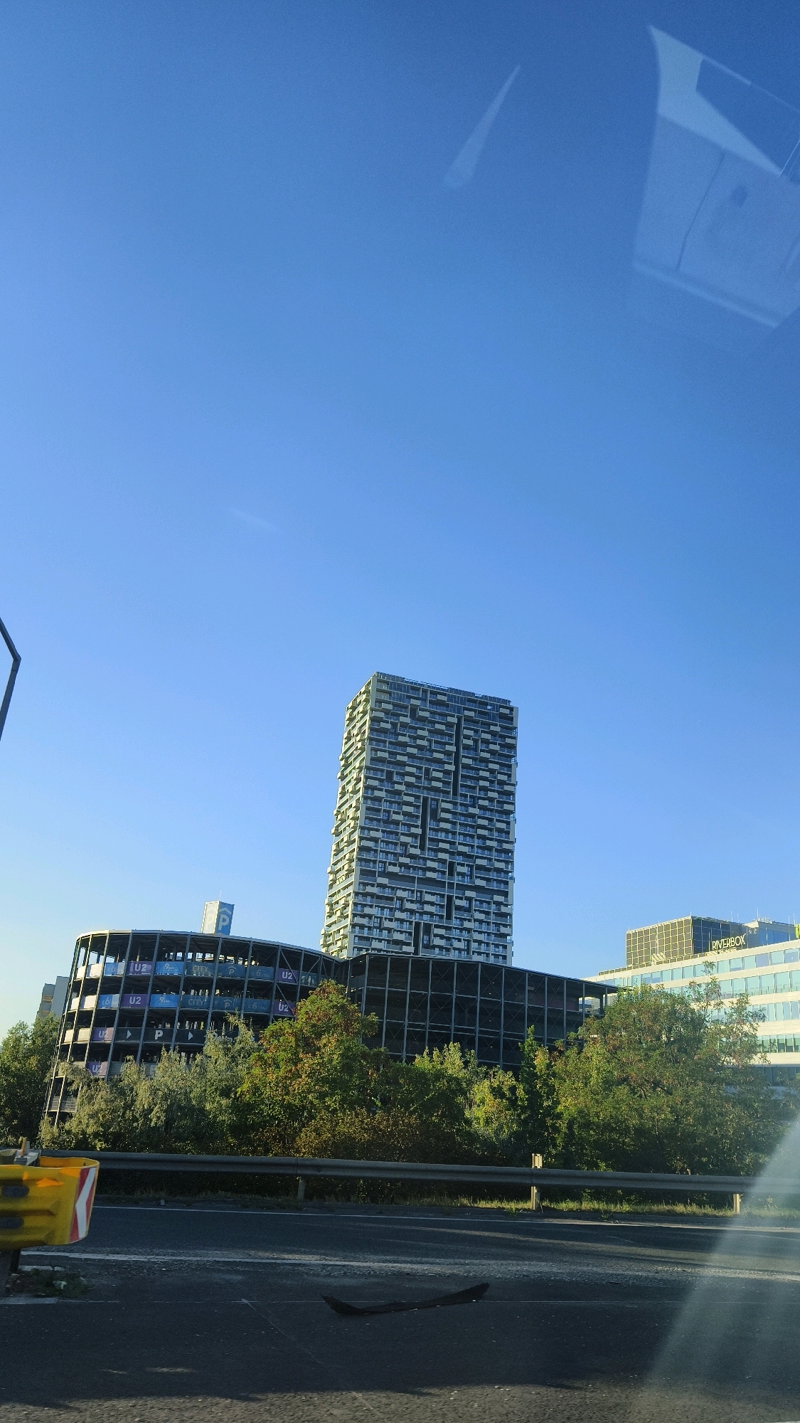 A tall, modern apartment building with a light-colored facade featuring many balconies stands against a clear blue sky. To its left, a multi-story, open-air parking garage with a curved design is visible, displaying signage for "U2" and "P" on its levels. Behind the parking garage, a lower, glass-fronted office building with the name "RIVERBOX" on its facade can be seen. The buildings are partially obscured by lush green trees in the foreground. A highway with a metal guardrail runs across the bottom of the image, with a yellow and white traffic barrier on the left side. The overall scene depicts an urban environment with a mix of residential, commercial, and infrastructural elements, likely taken from a vehicle on the highway.