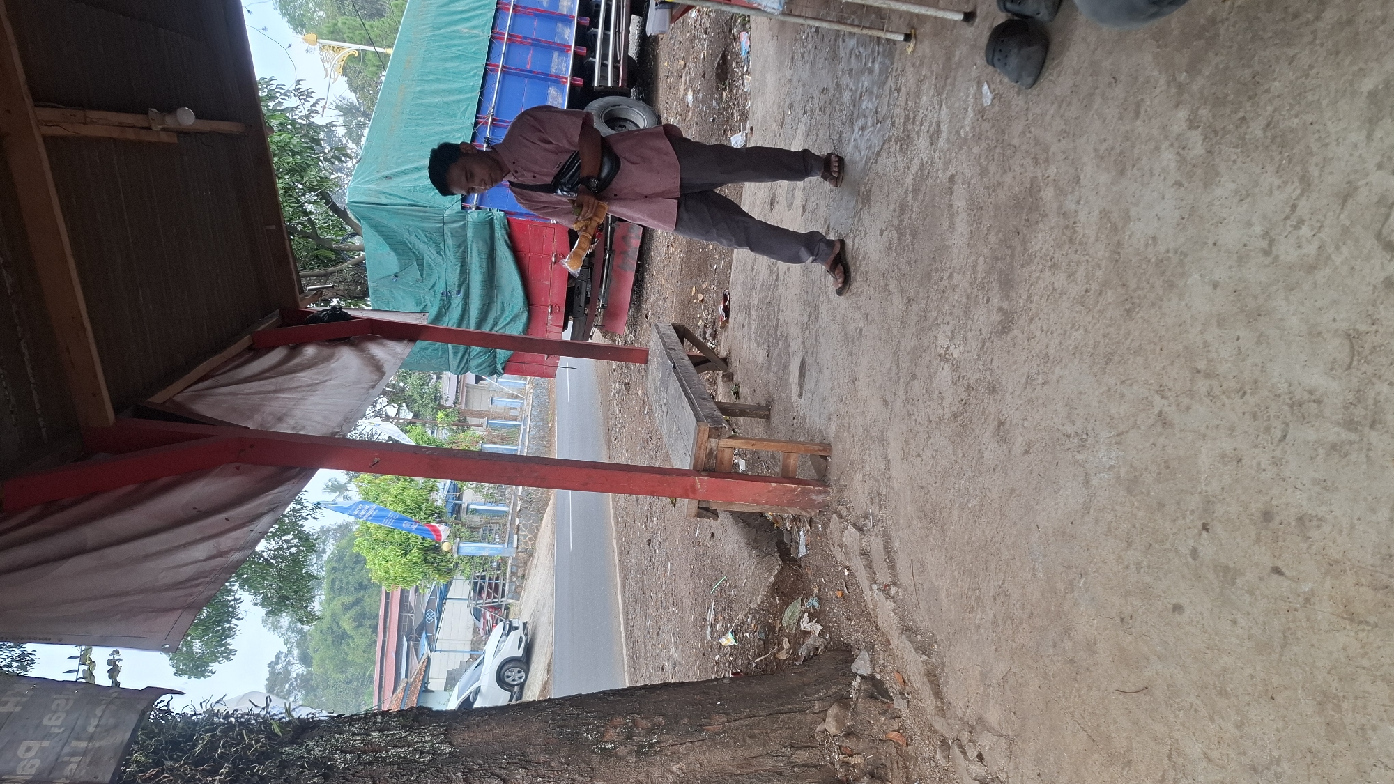 A young man in a pink collared shirt and grey pants stands on a concrete path, holding a snack. He is wearing a black fanny pack across his chest and brown sandals. To his left, a wooden bench with weathered planks sits on a dirt and gravel area. Behind the bench, a large truck with a green tarp covering its cargo is parked. In the background, a road runs parallel to the scene, with a white car parked on the side. Trees and buildings are visible further in the distance. The location is the side of a road in Cilaku, Indonesia, under a weathered shelter with red poles and a corrugated metal roof.