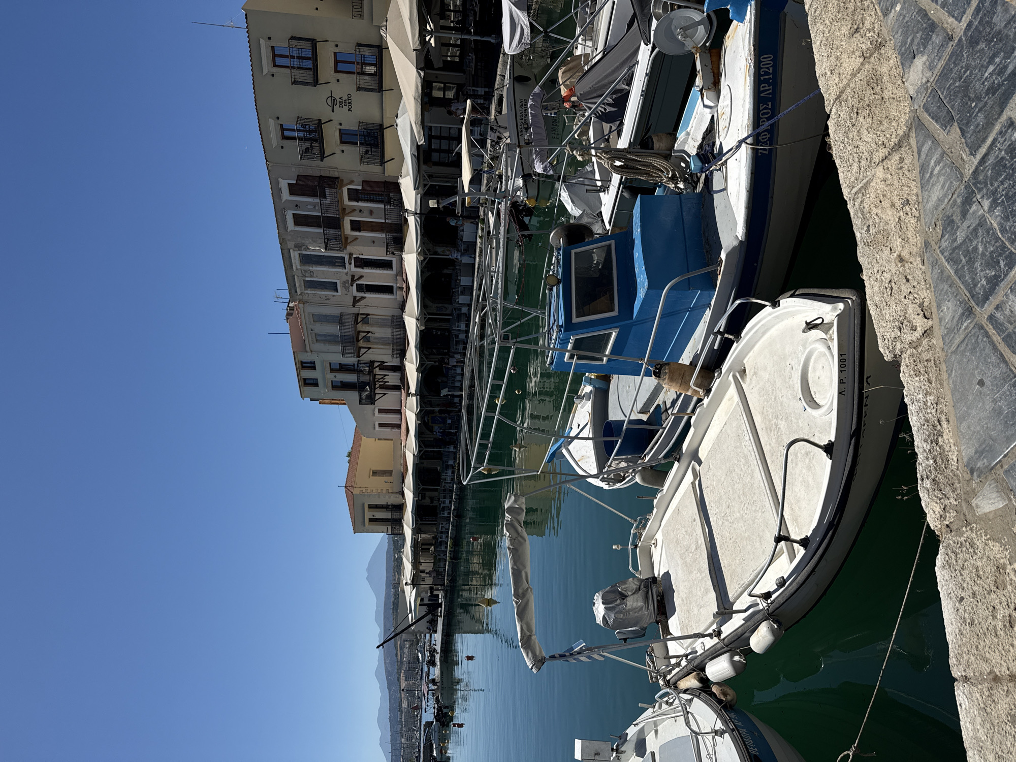 The image depicts the Venetian harbor in Rethymno, Greece. In the foreground, several small boats are moored. The closest boat on the right has a white hull with a blue stripe and appears to be a fishing boat, as indicated by the netting and ropes. It has the registration "ΖΕΦΥΡΟΣ ΛΡ. 1200" on its side. Next to it, to the left, is another white boat with a light-colored canopy. The leftmost boat in the immediate foreground is partially visible, showing its white hull.
In the mid-ground, more boats are docked, some with their covers on. Beyond the boats, a promenade is lined with buildings featuring arched windows and balconies. Several establishments with outdoor seating and umbrellas suggest a waterfront dining or cafe area. The architecture of the buildings is characteristic of Venetian influence, with pastel-colored facades and terracotta roofs. A prominent sign on one building reads "DEA DEL PORTO."
The background reveals a calm body of water reflecting the sky and buildings. In the distance, hills and mountains are visible under a clear, bright blue sky, suggesting a sunny day. The overall scene conveys a peaceful and picturesque maritime atmosphere in the historic city of Rethymno.