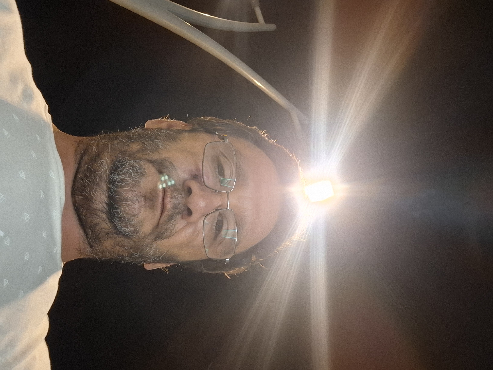 A low-angle, wide-angle selfie of a middle-aged man with glasses and a beard taken outdoors at night. The man is looking up at the camera, with a bright streetlight directly above him creating lens flare. The man is wearing a light-colored t-shirt with a subtle pattern. In the background, there are dark sky and some abstract structures that appear to be part of a street lamp. There are several small, bright green objects clustered on the man's upper lip and chin. The image quality is slightly grainy, possibly due to low light conditions. The location is Yerocham, Israel.