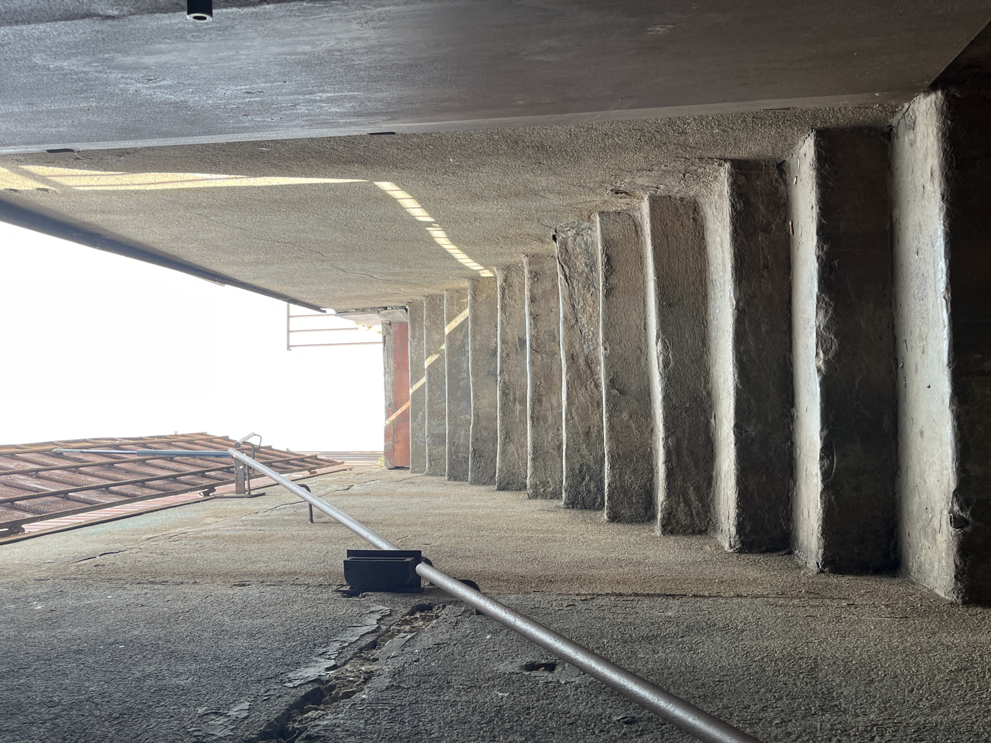 The image captures a narrow, concrete or stone passageway or underpass in Barcelona, Spain, viewed from a low angle looking upwards and forward. The ceiling, which is the underside of an elevated structure, is a flat expanse of grey concrete. Along the right side, a series of roughly textured, vertical concrete or stone steps or structural elements recede into the distance, creating a strong repetitive pattern. The floor of the passageway is a textured concrete surface, showing signs of cracks and wear. A metallic pipe or handrail extends diagonally from the lower right foreground towards the mid-left, supported by a dark, rectangular mount on the ground.
To the far left of the frame, a bright, blown-out opening indicates the exterior, where a section of a terracotta-tiled roof structure is visible. The scene is illuminated by strong, natural daylight streaming in from this opening, casting distinct shadows across the floor and highlighting the textures and forms of the concrete elements. The bright light and clear shadows suggest it is daytime on a sunny day. No people or specific activities are taking place. The overall impression is one of an aged, utilitarian architectural space, emphasizing geometric forms, material textures, and the interplay of light and shadow. There is no visible text in the image.