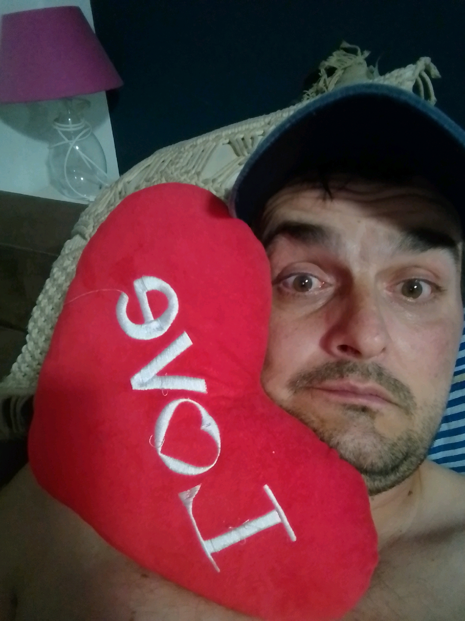 A man is shown holding a red heart-shaped pillow with the word "love" embroidered on it in white. He is wearing a dark blue baseball cap. The background appears to be a bedroom, with a lamp and what looks like a woven wall hanging visible. There are no clear indicators of the location in Wedel, Germany.