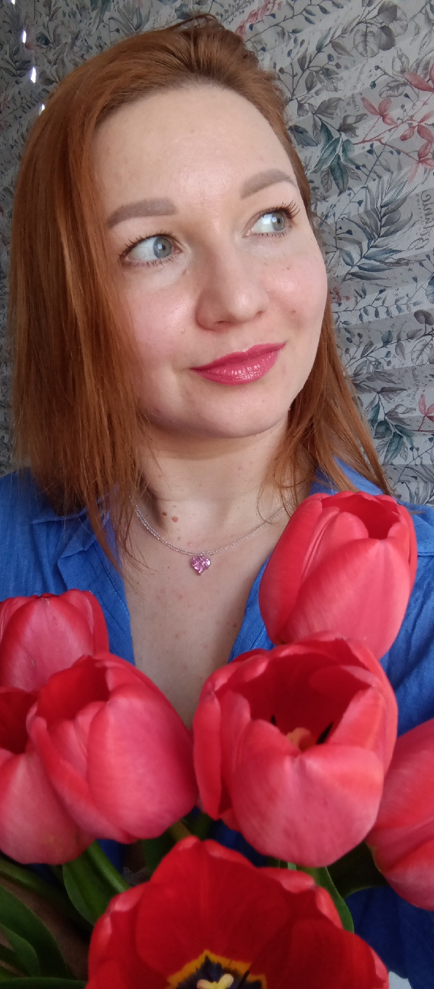 A woman with red hair and blue eyes is holding a bouquet of red tulips. She is wearing a blue shirt and a silver necklace with a pink heart pendant. The background is a floral patterned wallpaper. The location is Unknown, Unknown.