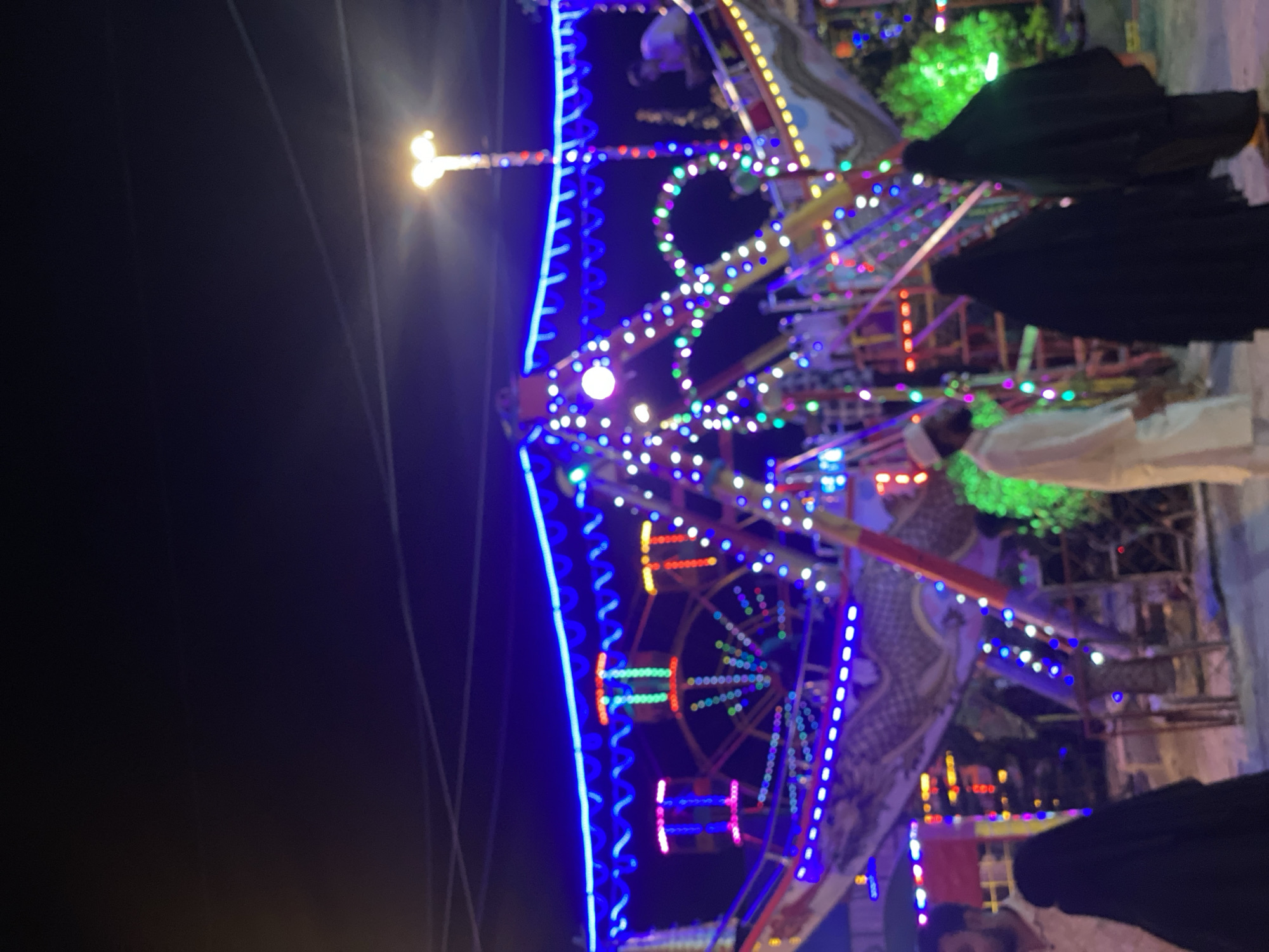 Here is a description of the image:
The image depicts an illuminated Ferris wheel at night. The Ferris wheel is adorned with multi-colored lights. In the foreground, there are people, some wearing hijabs. The setting appears to be an outdoor event, possibly in Dhaka, Bangladesh, based on the presence of individuals in traditional attire and typical urban lighting. The environment suggests a fair or festival, likely held in the evening.