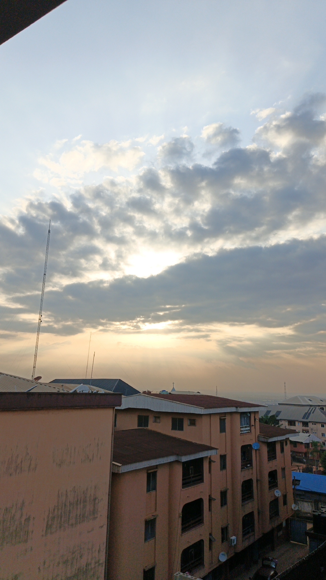 This image captures an urban landscape scene, likely at either sunrise or sunset, from an elevated viewpoint looking over buildings in Onitsha, Nigeria. The sky dominates the upper half of the frame, showcasing a mix of cumulus and stratocumulus clouds. Warm, golden sunlight dramatically breaks through the clouds, forming distinct crepuscular rays that spread across the sky and illuminate the atmospheric haze.
Below the horizon, a dense collection of buildings fills the lower portion of the image. The most prominent structures are multi-story buildings, likely residential apartments, characterized by their peachy or salmon-pink facades and dark brown or terracotta-colored sloped roofs. Many of these buildings feature balconies with dark railings and visible windows. Several white satellite dishes are affixed to the exteriors of these buildings. A large, plain wall of a similar hue occupies the immediate left foreground.
A very tall, slender communication or broadcast tower stands prominently on the left side, extending high into the sky, equipped with various antennas and components. Another, shorter antenna is visible closer to the horizon on the left. In the mid-ground, a white cross can be seen atop a distant building, possibly indicating a church or religious institution. Further into the distance, more buildings with varied roof colors (including blue and grey) are visible, along with another faint, tall structure on the right horizon. No people or specific activities are discernible. The overall impression is one of a peaceful urban expanse bathed in the soft, diffused light of dawn or dusk.