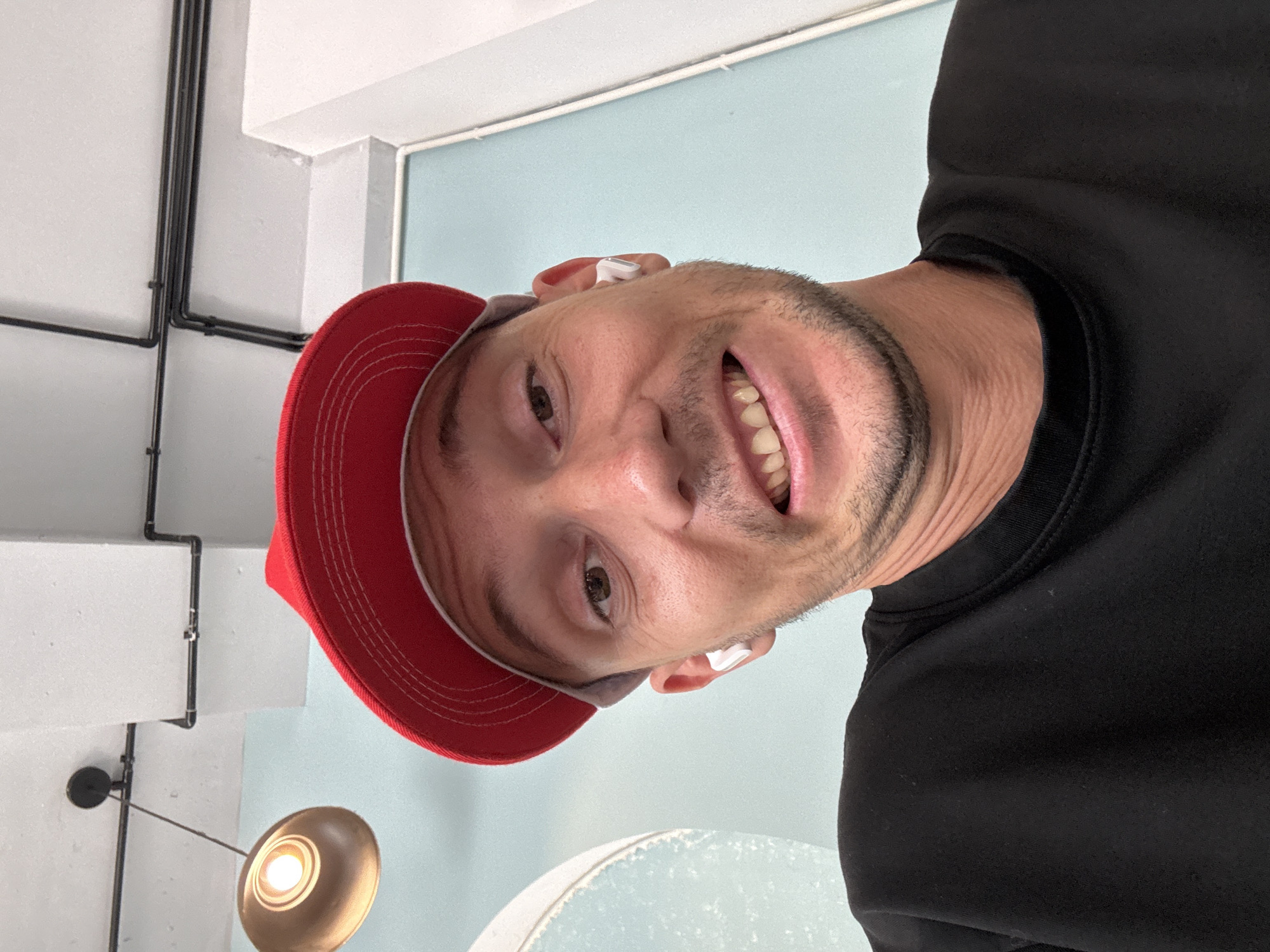 A man is positioned looking down at the camera at an angle. He has a red baseball cap with a white border. He is wearing in-ear headphones. He is wearing a black long sleeved shirt. The background is comprised of white ceiling and wall fixtures, a light blue wall, and a hanging light fixture. There is no visible information to suggest the location of the image is Nha Trang, Vietnam.