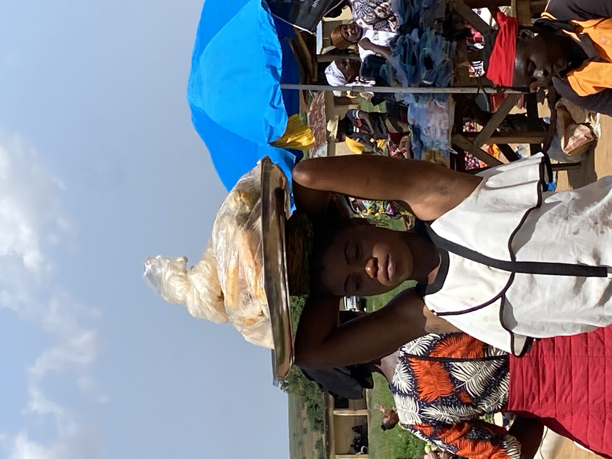 In a city most likely Riyom, Nigeria, a woman is carrying a tray of food on her head. The food is in a plastic bag. She is wearing a white shirt with black trim and a red and patterned skirt. In the background, there are people selling clothes and goods under a blue umbrella. The sky is blue with some clouds.