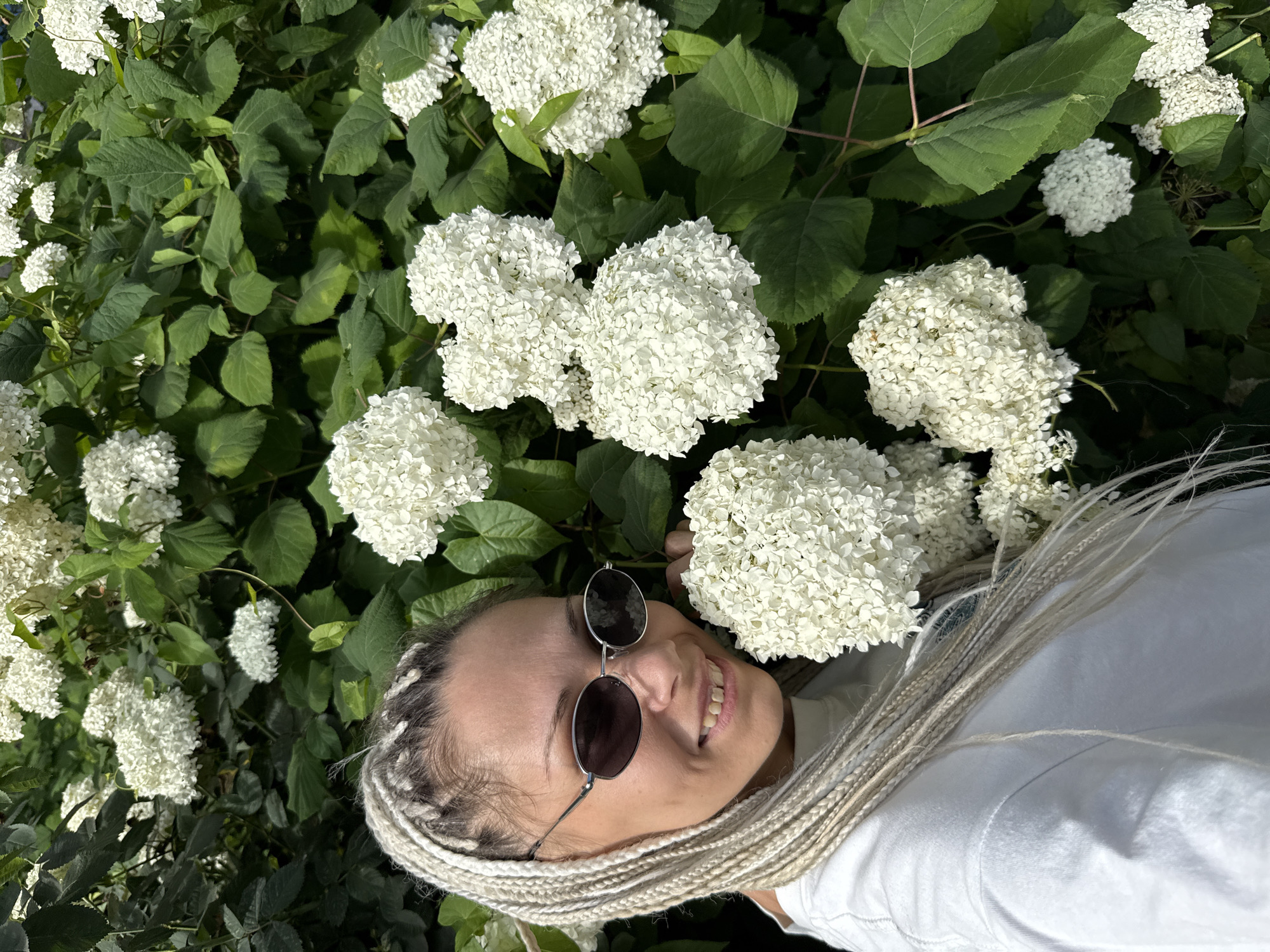 An outdoor medium shot shows a white woman with blonde dreadlocks wearing sunglasses and a white t-shirt. She is smiling and looking to her left. She is positioned in front of a bush with many white flowers and green leaves. The image is lit by natural sunlight.