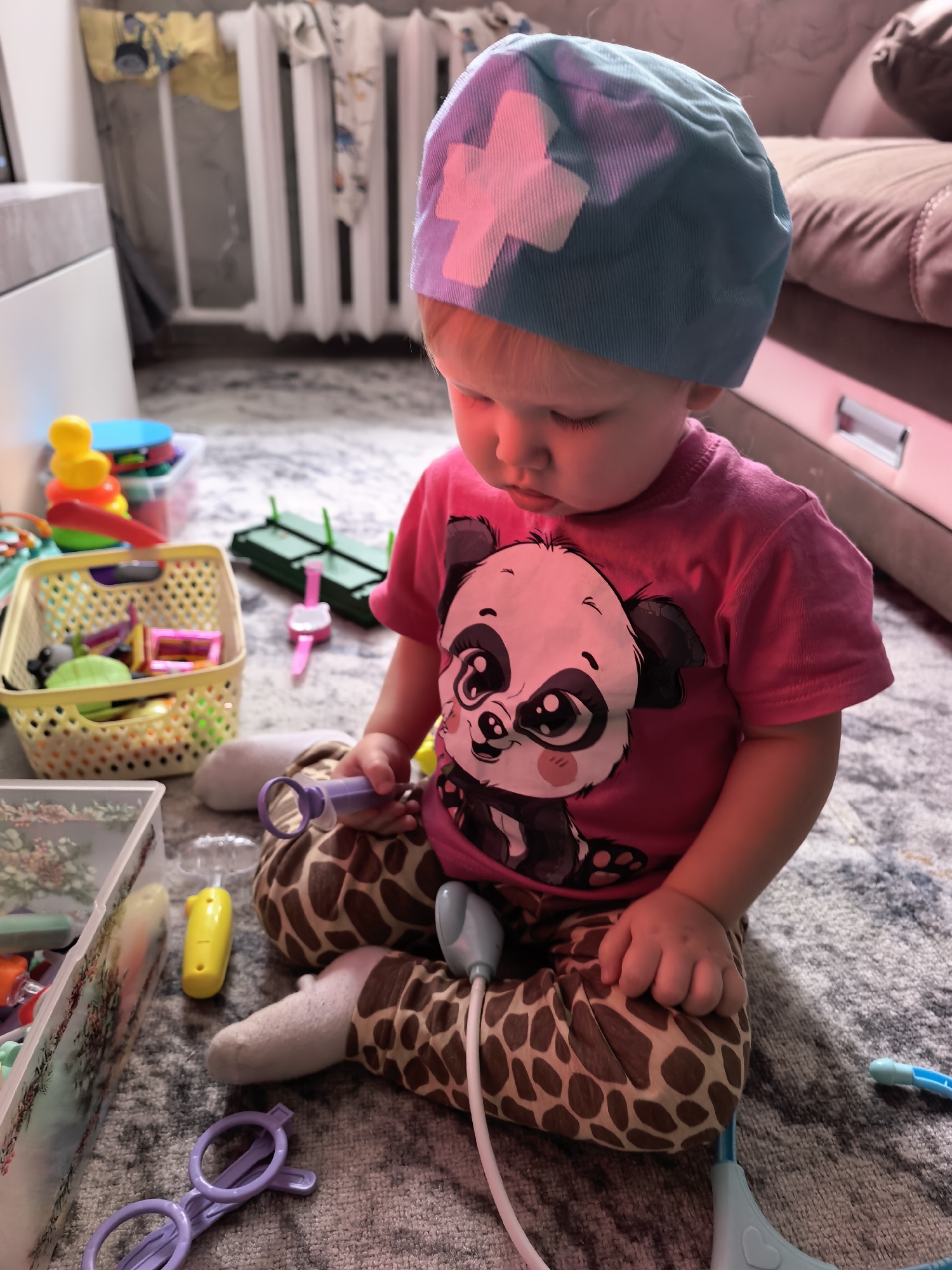 A young child, likely under the age of three, is sitting on a patterned rug in what appears to be a home environment. The child is wearing a pink t-shirt with a panda graphic and giraffe-patterned pants, and a blue surgical cap with a white cross. The child is holding a purple toy stethoscope and appears to be playing with doctor's kit toys scattered around. Several toy containers and other children's items are visible in the background, including a radiator and a sofa. The lighting suggests an indoor setting, possibly during daytime. No specific landmarks or elements definitively place the scene in Sterlitamak, Russia.
