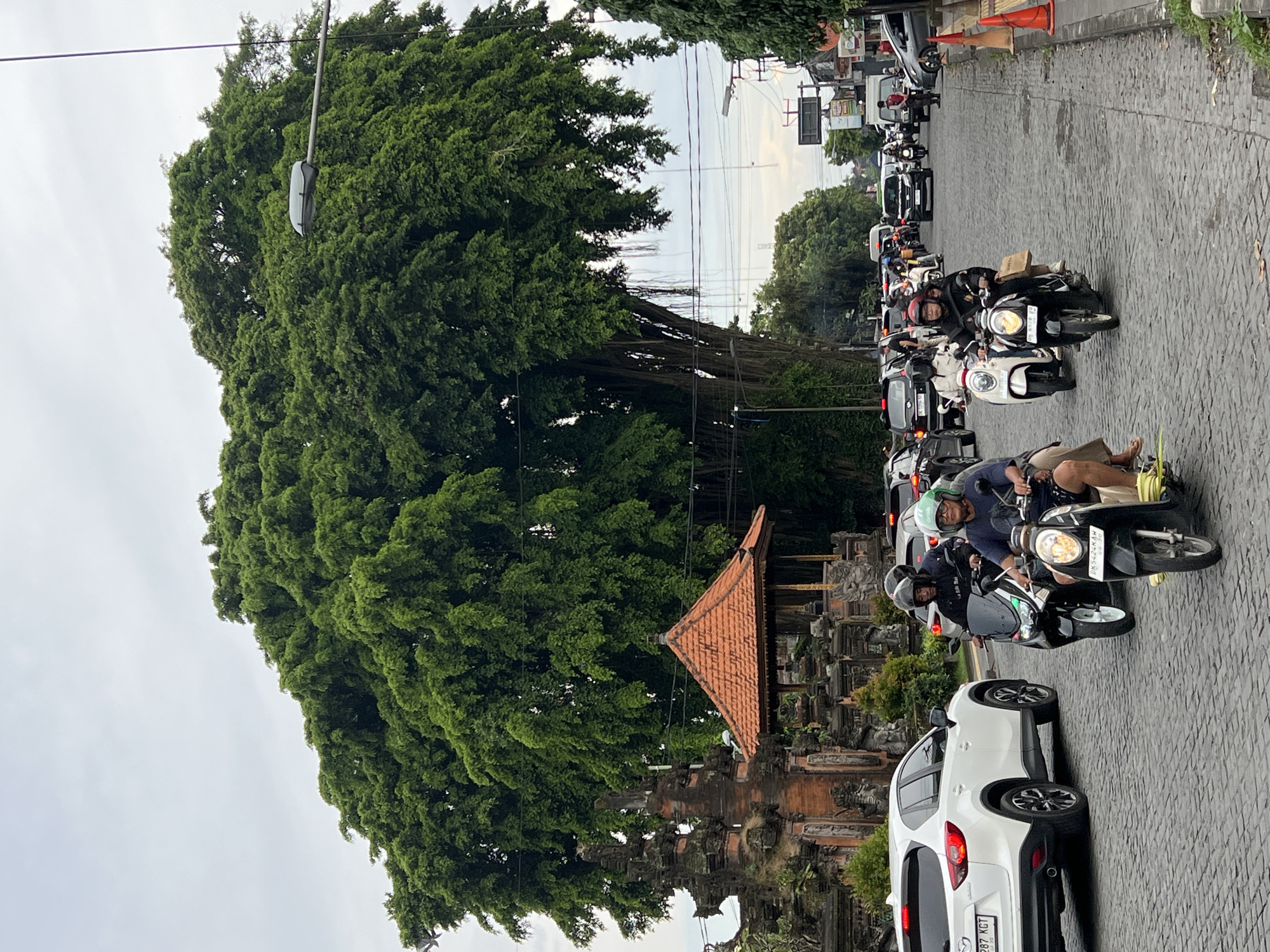 The image depicts a street scene in Peliatan, Indonesia. A massive Banyan tree dominates the left side of the frame, its foliage obscuring a portion of a traditional Balinese gate structure with a tiled roof. The street is paved with cobblestones and filled with a traffic jam of cars and scooters. Several people are riding scooters, some wearing helmets. A white car is visible in the foreground. Overhead, power lines stretch across the sky. The sky is overcast.