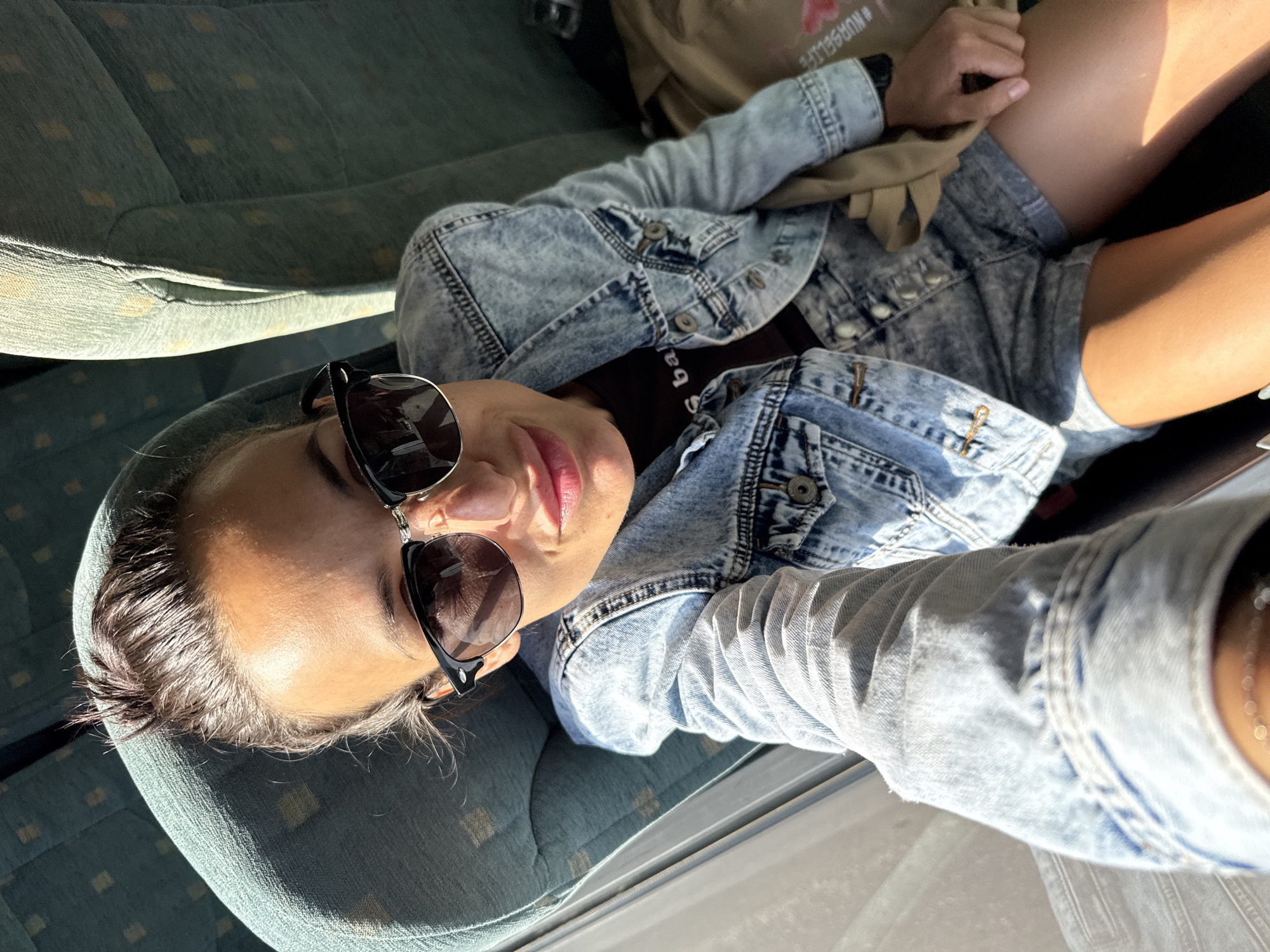 A woman wearing a denim jacket, black shirt, and denim shorts reclines on a blue patterned seat. She is also wearing sunglasses. A backpack is visible near her head. The location appears to be inside a vehicle, possibly a bus, likely within a city, based on surrounding infrastructure. The exact location within Pleven, Bulgaria, is not determinable from the image.