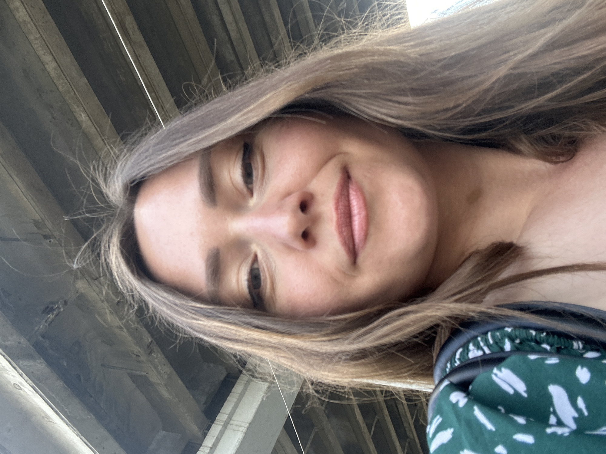 The image is a selfie taken from a low angle, showing a woman's face from the chin up. She has light brown hair with blonde highlights that is somewhat windswept. Her eyes are open, and she is smiling slightly. She is wearing a green dress with a white abstract pattern. A dark strap, possibly from a bag, is visible on her shoulder. The background appears to be the underside of an overpass or bridge, with concrete structures and exposed rebar. The lighting suggests it is daytime, and the overall impression is casual and outdoors. The location of Bila Tserkva, Ukraine, is not evident from the visual information in the image.