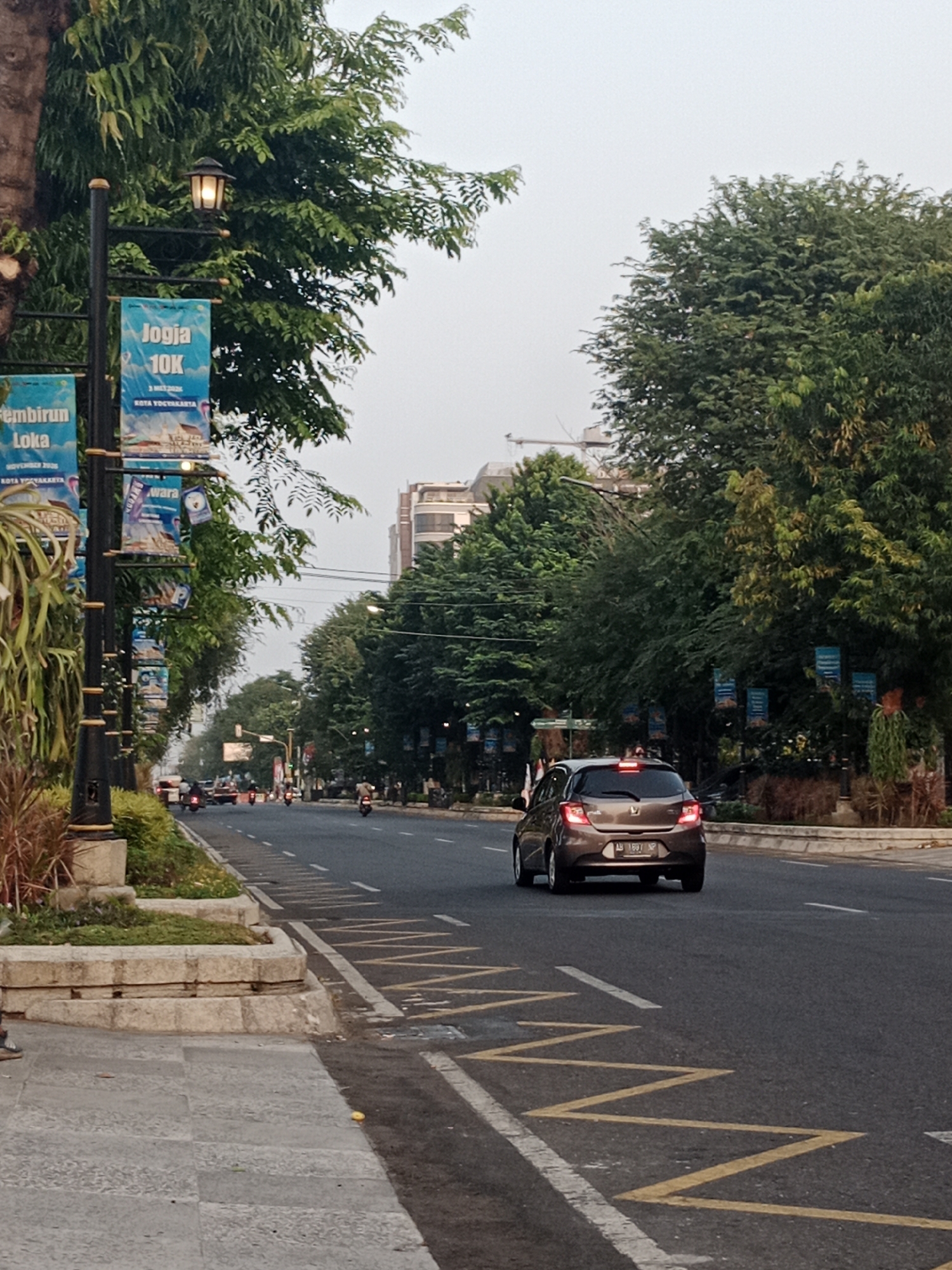 The image depicts a street scene in the city of Yogyakarta, Indonesia. On the left side of the road, there are lampposts with banners attached. One banner reads "Jogja 10K". Further down the street, a brown car is driving away from the camera. The road is asphalt and has yellow and white markings. The environment is lined with trees, and buildings can be seen in the background. The sky is overcast, suggesting it may be early morning or late afternoon.
