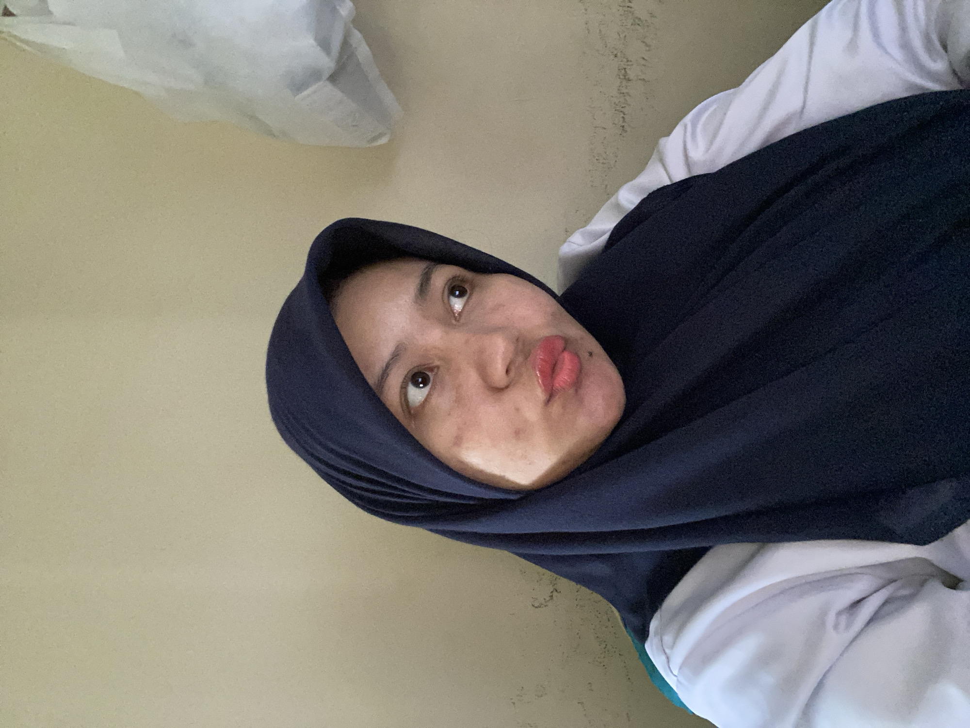 A woman wearing a navy blue hijab and a white top is pictured from the chest up, looking to her right. Her lips are pursed, and she has light-colored skin with some blemishes on her cheeks. The background is a plain, light-colored wall. To the right of the woman, a white plastic bag is hanging on the wall. The image appears to be a selfie taken indoors. This scene is located in the city of Banjarbaru, Indonesia.