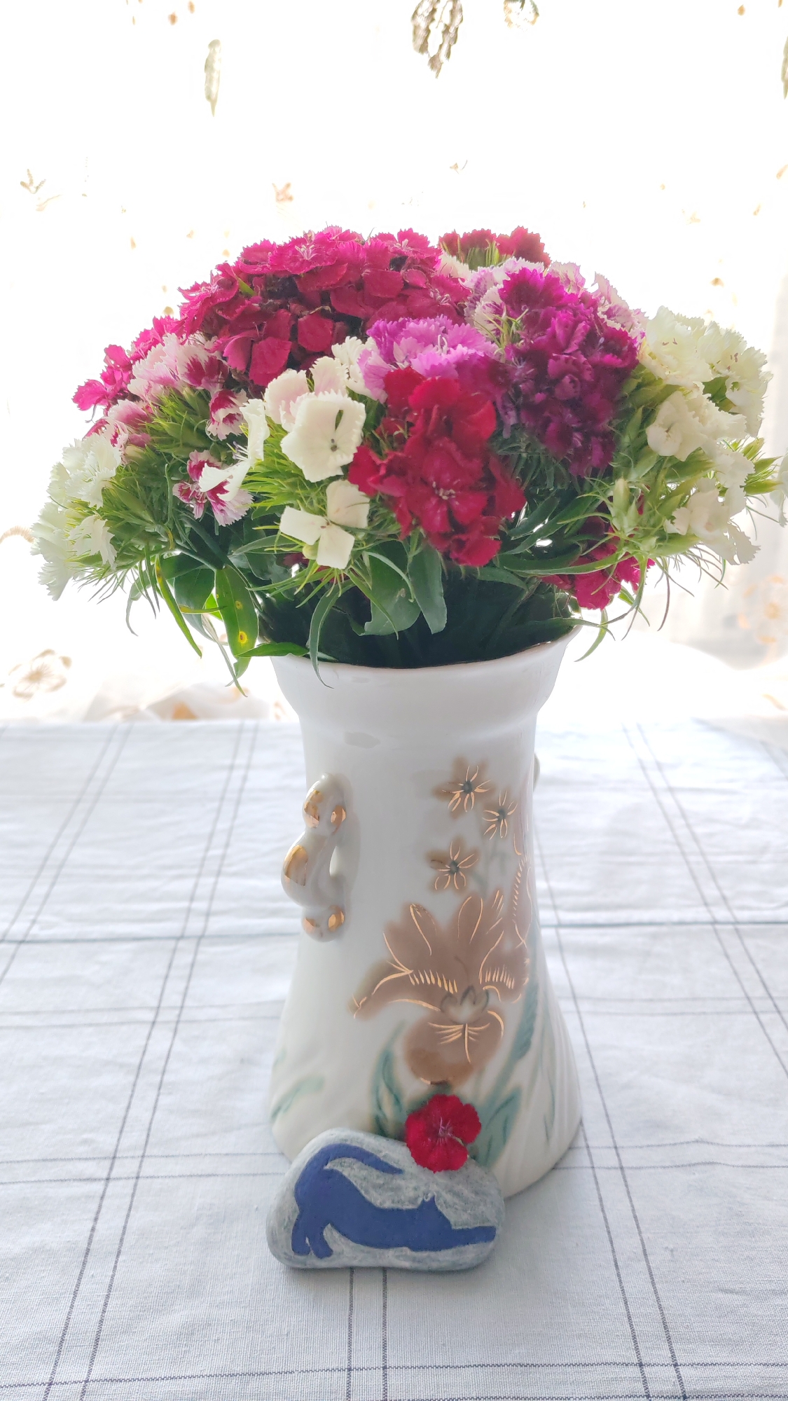 A bouquet of dianthus flowers in shades of white, pink, and deep red is displayed in a white porcelain vase with gold floral accents. The vase is adorned with a small, gold figurine of what appears to be a monkey or a similar creature on its left side. In front of the vase, resting on a light grey and white checkered tablecloth, is a flat, grey stone decorated with a blue silhouette of a cat in a stretching pose. A single small red flower petal sits atop the stone, next to the cat silhouette. The background consists of a sheer, white curtain with subtle, light brown floral patterns, allowing diffused daylight to illuminate the scene.
