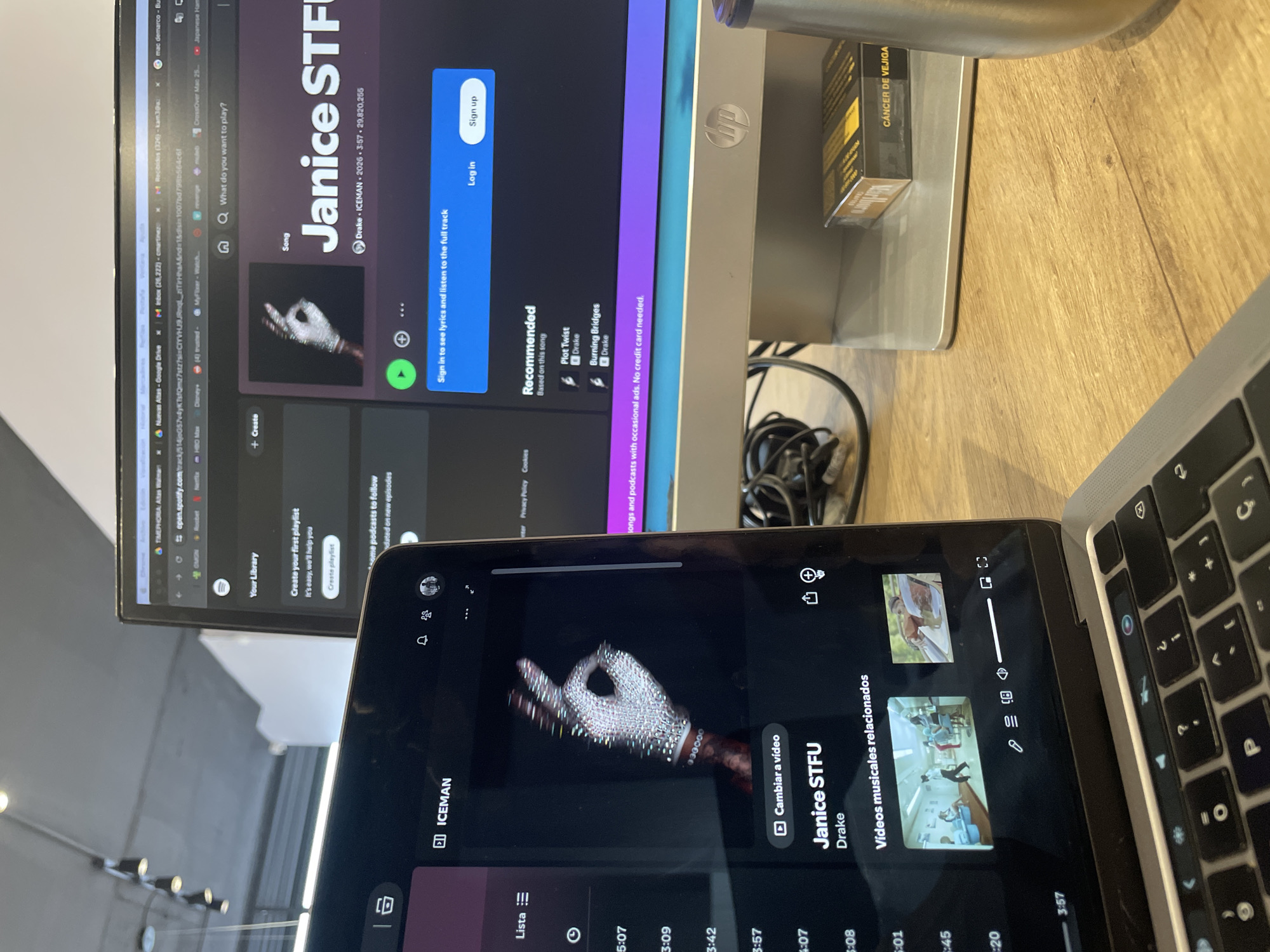 A cluttered workstation is depicted in the image, likely indoors. An HP monitor dominates the scene, displaying the Spotify application open to the song "Janice STFU" by Drake. The same song is displayed on a mobile device in the foreground. A silver MacBook is adjacent to the mobile device. A box of "CÁNCER DE VEJIGA" is seen by the computer.
No specific location is discernible from the image. Based on the box of medicine labelled “CÁNCER DE VEJIGA” (bladder cancer) in Spanish, the location is somewhere in a Spanish-speaking country. Given other clues, it is most likely the city of Mexico City, Mexico.