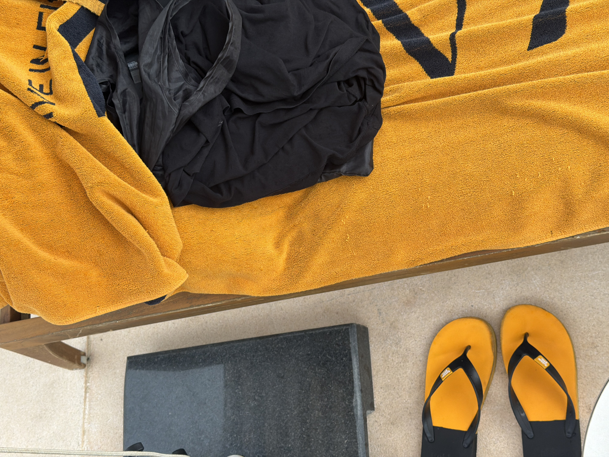 The scene presents a top-down view of a lounge chair, likely poolside, in Choeng Thale, Thailand. The lounge chair is covered with a yellow towel with dark blue stripes and lettering along the top left. A crumpled black garment rests on the towel. Below the chair, yellow flip-flops with black straps are placed on the ground next to a gray stone or concrete slab. The ground is light beige. The image is brightly lit, suggesting a sunny day. No people are visible.