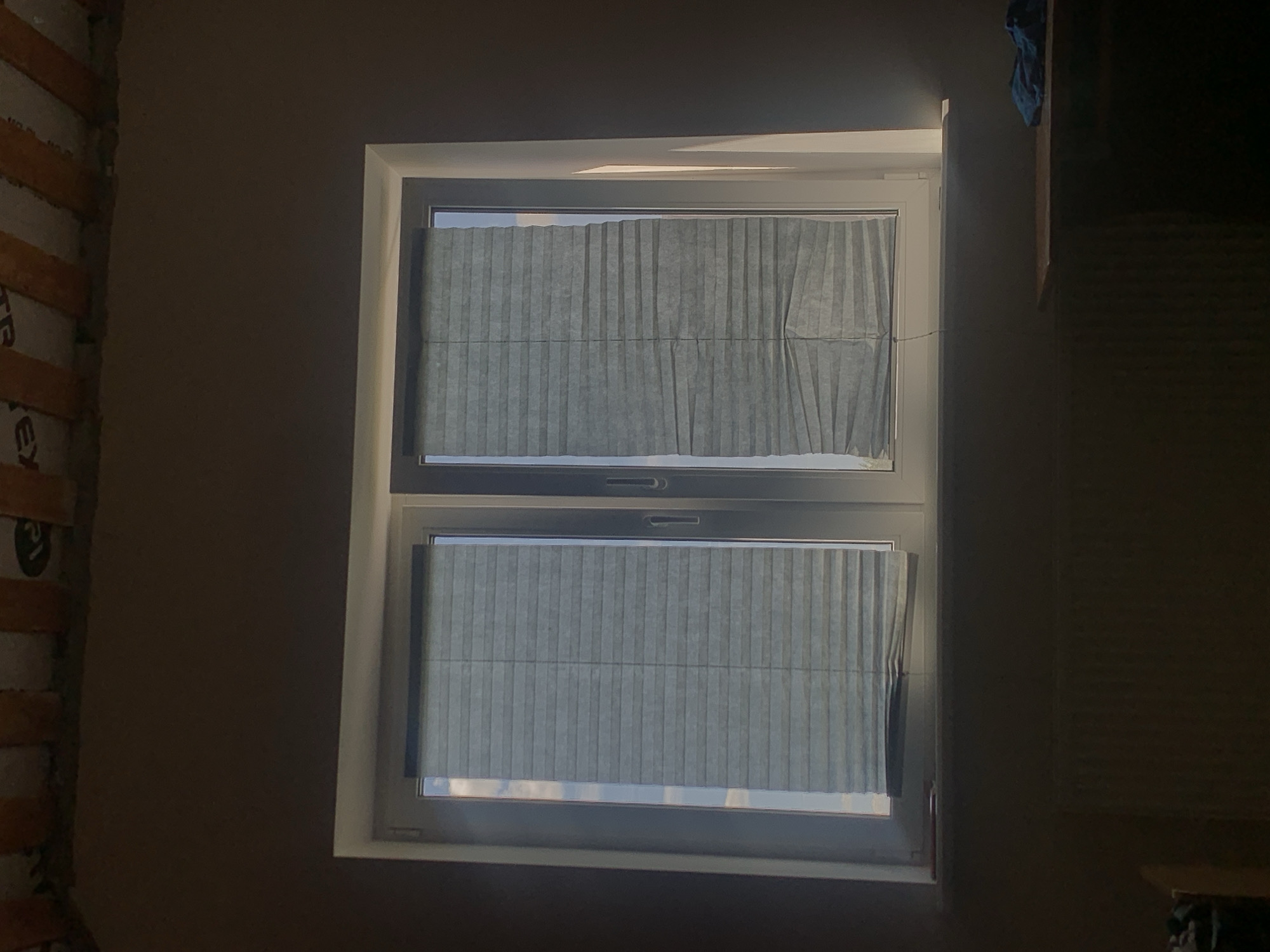 The image depicts an interior view of a room with a window. The window is a double-paned unit with light gray pleated blinds covering the glass. The left blind is partially lowered, while the right blind is almost fully lowered, with its bottom edge creased. Sunlight is visible streaming in from the left side of the window, casting shadows on the right side of the wall. Above the window, exposed wooden beams and insulation material are visible, suggesting construction or renovation is in progress. The walls are painted a dark, muted color. Below the window, a radiator is present, and to the right, a portion of a wooden surface is visible with a blue cloth draped over it. The overall lighting is dim, with the sunlight creating a strong contrast. The scene is devoid of people or any overt activity. The image does not contain any elements that can identify the location as Minsk, Belarus.