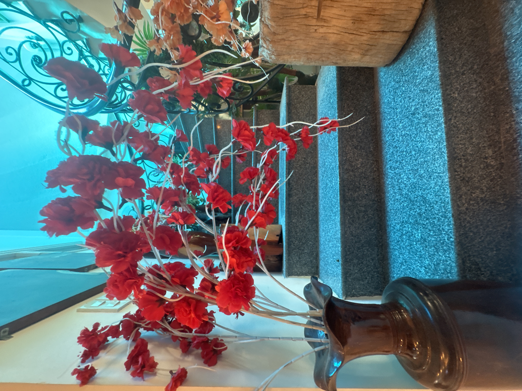 In Bac Nha Trang Ward, Vietnam, a dark brown ceramic vase with a flared rim stands on the lower left, holding a large arrangement of artificial red flowers with white stems. The flowers spill upwards and to the right, some hanging down. Behind the vase and flowers, a staircase with gray, speckled treads and risers ascends towards the upper right of the frame. The stairs are illuminated by a vibrant, artificial blue light source, casting strong shadows. To the right of the stairs, a rough-hewn wooden log or stump is visible. In the background, a black wrought-iron railing curves upwards along the side of the stairs. The overall environment suggests an indoor setting, possibly a hotel lobby or a decorative entryway, with a focus on floral and staircase elements enhanced by dramatic lighting.