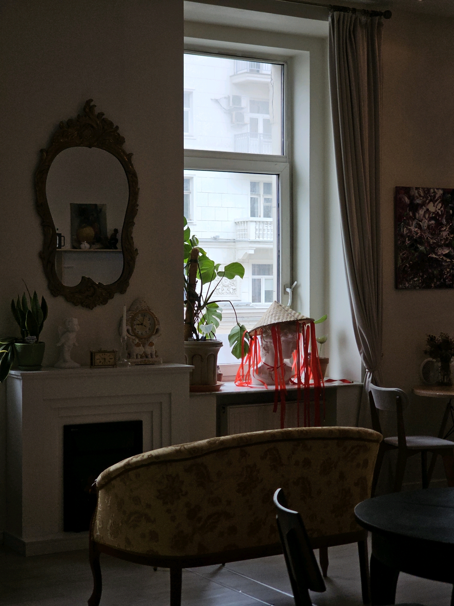 The image shows an interior scene, possibly a cafe or a room in a residential building in Moscow, Russia. The lighting is dim, with natural light coming from a large window. In the foreground, a vintage-style sofa with a floral pattern is visible. To the left, a white fireplace with decorative molding is present, with a black opening in the center. On the mantelpiece, there are several decorative items, including a plant in a green pot, a white figurine, a small rectangular clock, and an ornate clock. Above the fireplace, a large, ornate gold mirror hangs on the wall.
The window in the background reveals a view of buildings outside, suggesting an urban setting. On the windowsill, a plaster bust is adorned with a conical straw hat and red ribbons. Next to it, there is a potted plant. To the right of the window, a beige curtain is drawn back. Further to the right, a dark abstract painting hangs on the wall. In the mid-ground, a chair with a light-colored seat and dark legs is positioned near a dark round table. Another chair is visible further to the right. The flooring appears to be a dark surface, possibly wood or tile. The overall atmosphere is dimly lit and somewhat eclectic.