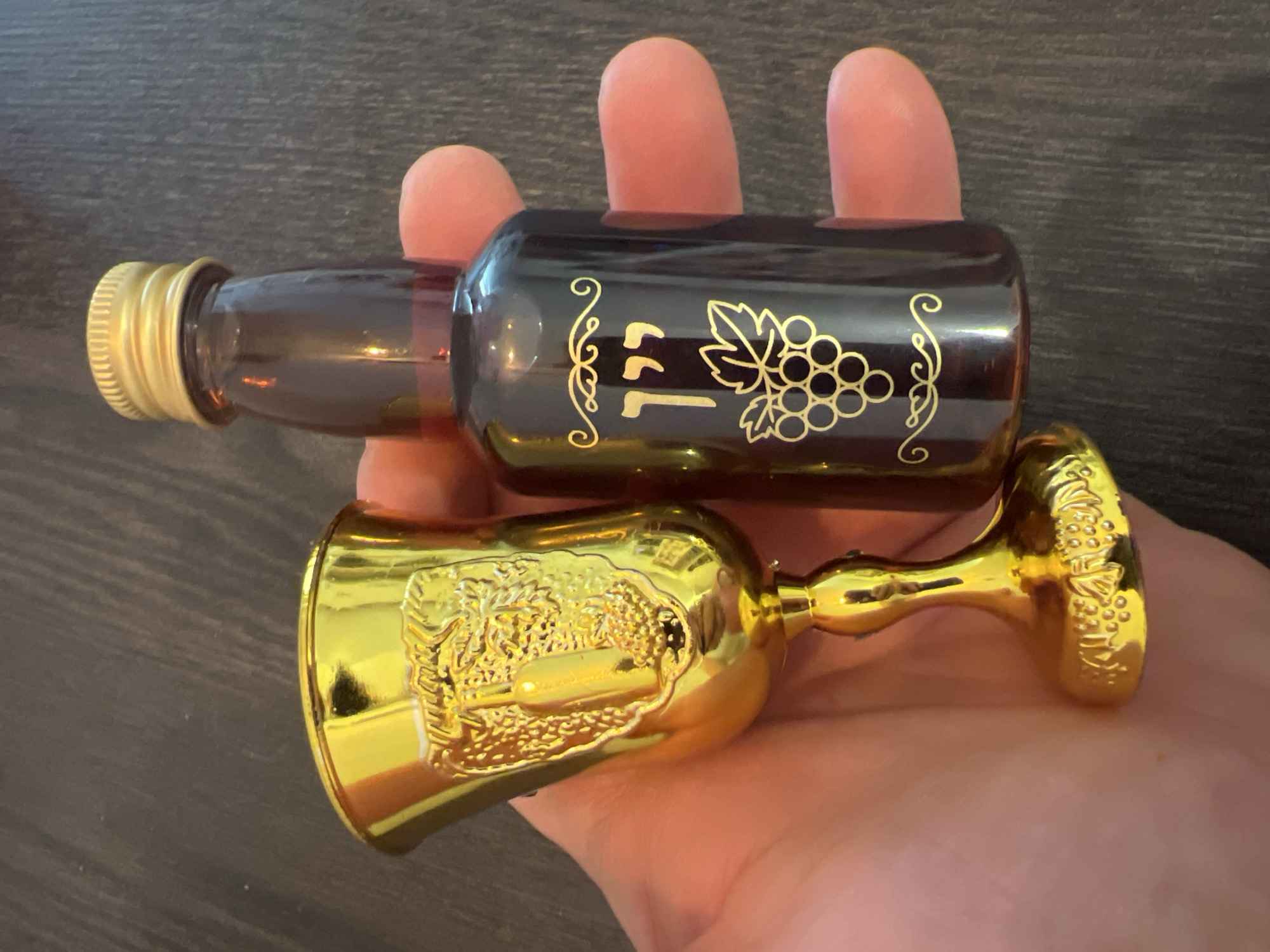 A light-skinned hand holds two decorative objects against a dark, wood-grained background. The primary objects are a small, dark amber-colored glass bottle and an ornate golden goblet.
The bottle contains a dark reddish-brown liquid and is sealed with a golden, ridged screw cap. Its side is adorned with golden printed designs, including a stylized vine with a cluster of grapes and leaves. To the left of the grapes, there are stylized characters that appear to be a '1' followed by what resemble the Hebrew letters 'ל' (Lamed) and 'ר' (Resh).
Below the bottle, a small golden goblet is held. The goblet features intricate embossed patterns on its cup, which seem to depict a crest or a detailed scene with figures. Its base also has a repetitive, textured design.
The scene is indoors, with no specific activity beyond the presentation of these items. No time of day, weather, or discernible emotions are indicated. The location context for this image is Ramat Gan, Israel.
