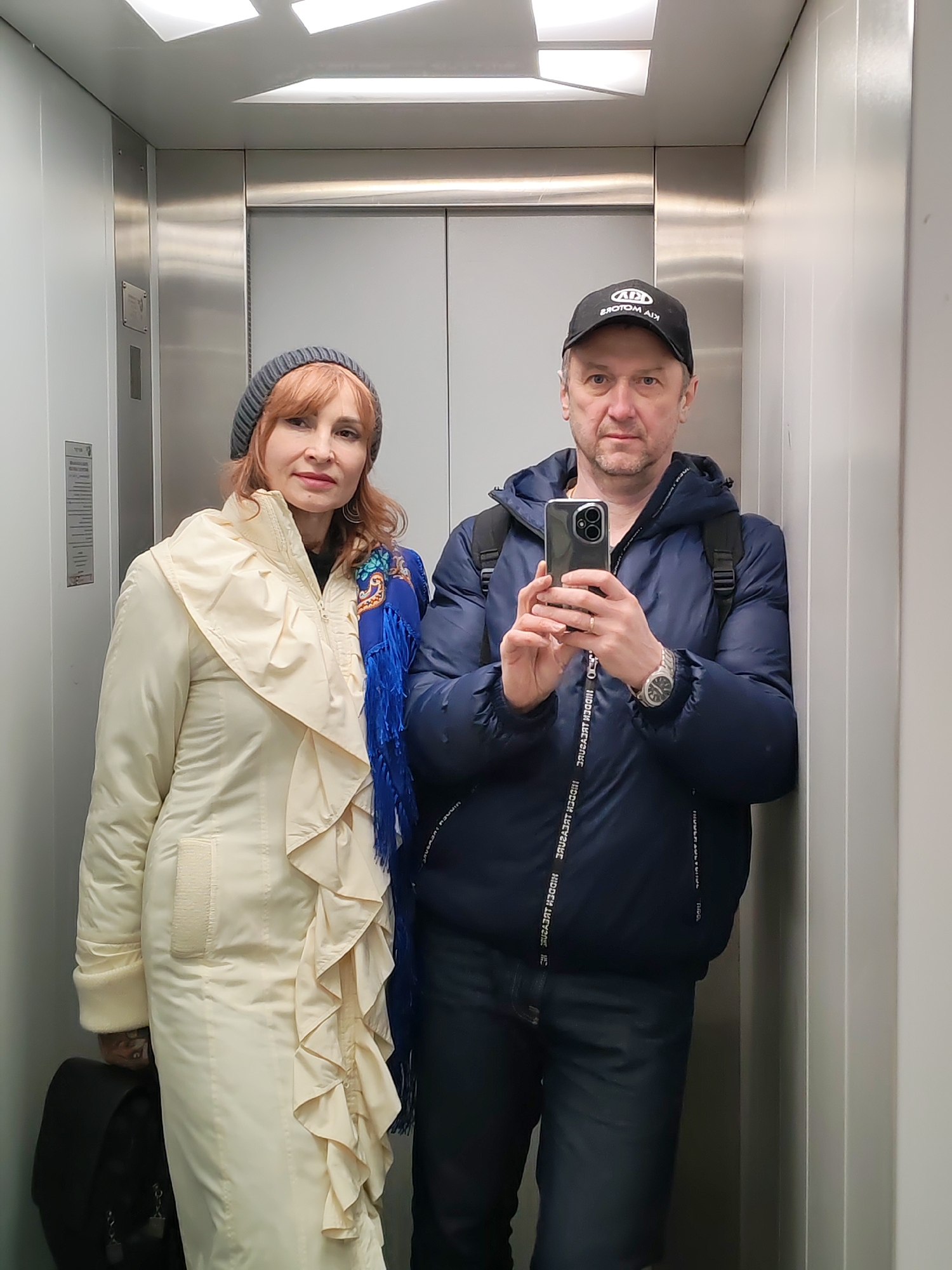 Two people are posing for a selfie in what appears to be an elevator. The woman on the left wears a long, light-colored coat with ruffles and a gray knitted hat, and carries a black bag. The man on the right wears a dark blue jacket, a baseball cap, and a watch. He is holding a smartphone in front of him to take the picture. They are surrounded by metal and white wall panels. A search reveals that this picture was taken in the city of Chaikovsky, Russia.