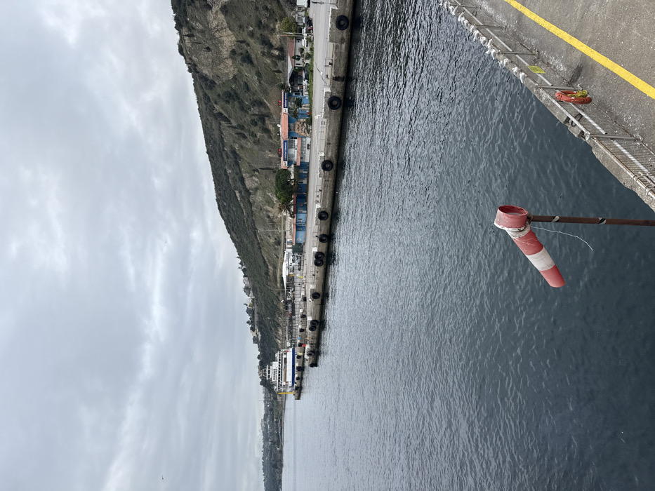 A harbor scene under a cloudy sky. A mountain rises along the left side of the frame, alongside a concrete pier with tires attached. The calm, dark water fills the right half of the image. A striped windsock is attached to a pole, indicating a light breeze. A yellow line marks the edge of the pier, with metal railings and ladders present. A large ferry is docked further along the pier, near buildings and stacked containers.