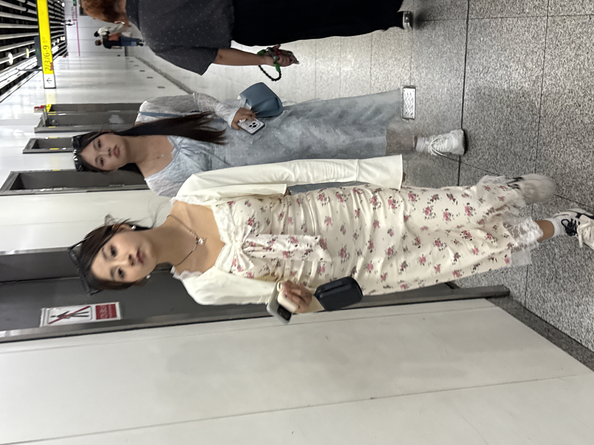 The image depicts two young women standing in what appears to be a subway station in the Yuzhong District of China. The women are in the foreground, facing the camera but with their bodies turned sideways. The woman closest to the camera wears a white floral dress with a matching white jacket and white sneakers. She is holding a phone and a small, dark-colored bag. The other woman is wearing a light blue dress and white sneakers, carrying a small blue purse and holding a phone. Behind them, other people walk along the subway platform. A yellow sign with Chinese characters is visible on a pillar on the left, and the platform has a gray tile floor.