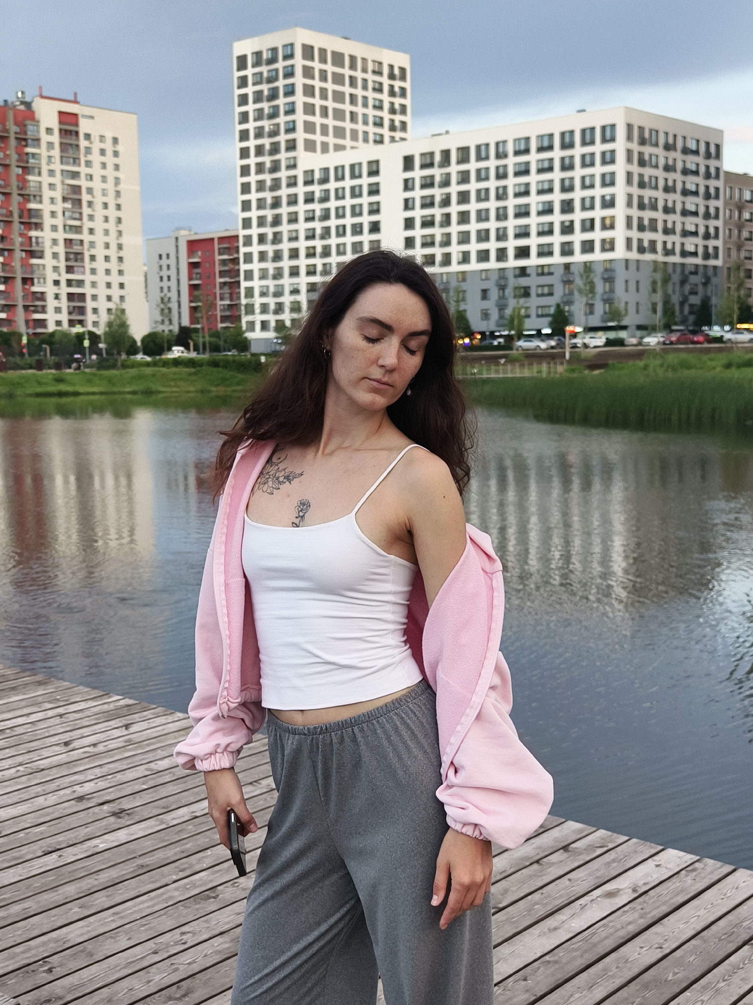A young woman with long dark hair, wearing a white tank top, a pink zip-up hoodie, and gray sweatpants, stands on a wooden boardwalk. She is holding a smartphone in her right hand. Her eyes are closed, and her head is tilted downwards. Behind her, a calm body of water reflects the sky and the surrounding modern buildings, which appear to be in a city park or a residential area. The buildings are tall, with white and red facades, and numerous windows. The sky is overcast with a hint of dusk. The overall scene suggests an urban setting, possibly in Tyumen, Russia, given the architectural style of the buildings and the presence of a park-like area with water.