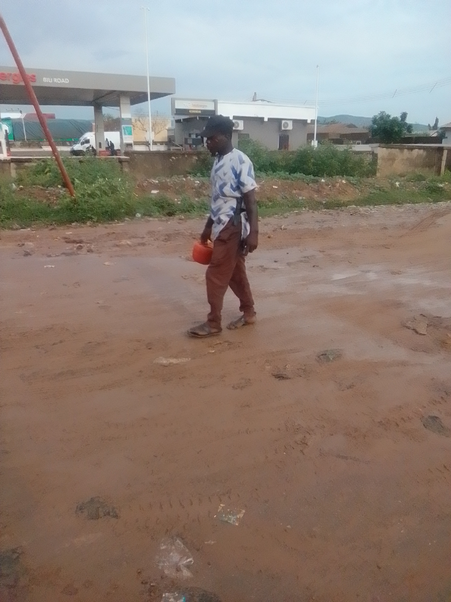 A man, appearing to be of African descent, walks along a muddy, unpaved road in Gombe, Nigeria. He is dressed in a blue and white patterned short-sleeved shirt and brown trousers. He carries a small orange container in his right hand, holding it close to his body. The man is walking from left to right in the frame, his gaze directed forward.
The environment is characterized by a reddish-brown, muddy terrain, indicating recent rainfall or damp conditions. Tire tracks are visible in the mud, suggesting vehicle passage. Scattered debris, including plastic bags, is present on the ground. In the background, a gas station with a prominent canopy and signage reading "ERGOS BIU ROAD" is visible. Beyond the gas station, a low-rise building with air conditioning units can be seen, and further in the distance, hills and vegetation are present under an overcast sky. The overall scene depicts an outdoor environment in a developing urban or peri-urban area.