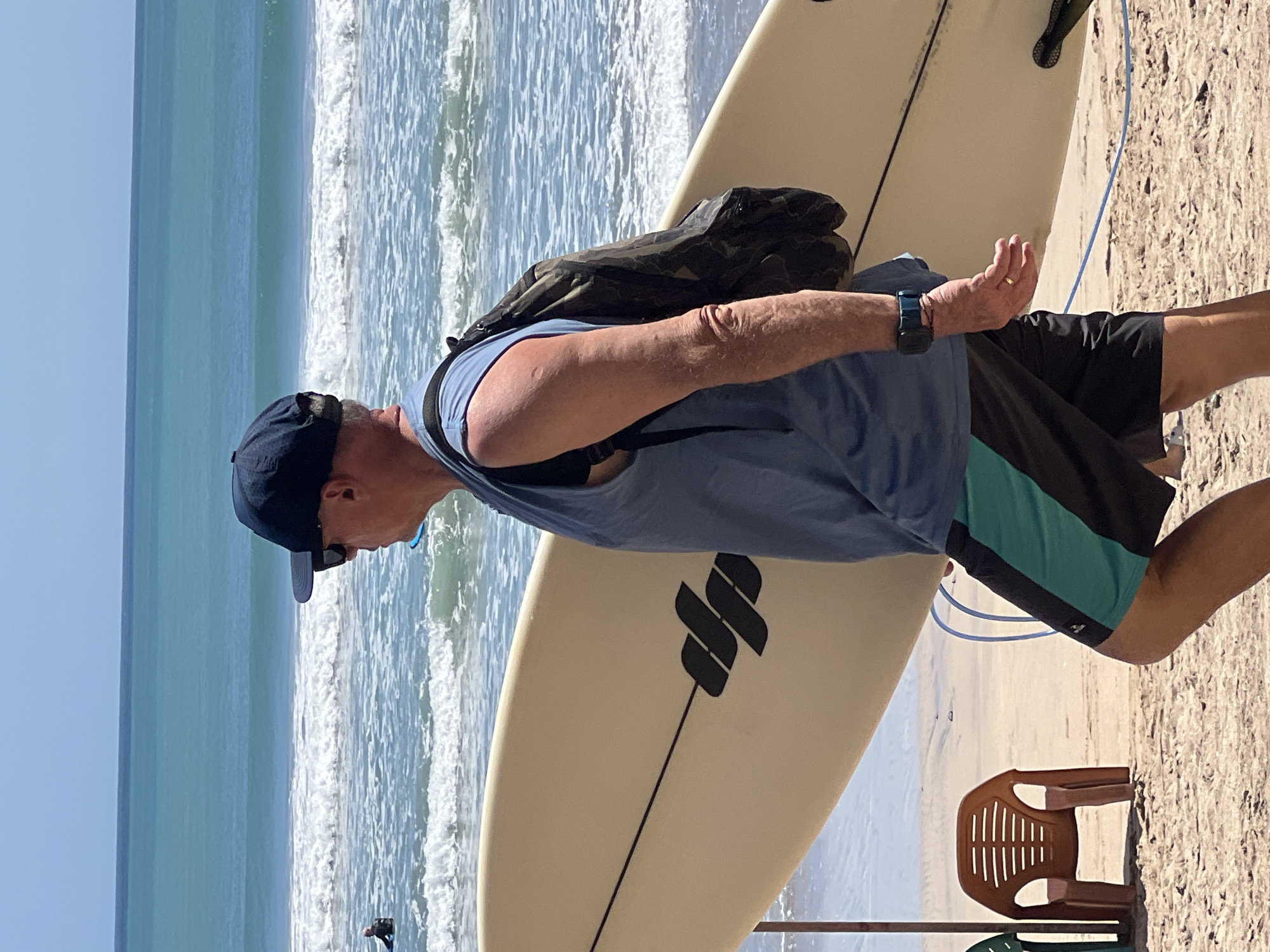 An older Caucasian male, wearing a blue sleeveless shirt, black shorts with turquoise accents, a dark baseball cap, and sunglasses, walks across a sandy beach in Kuta, Indonesia, carrying a pale yellow surfboard under his right arm. He has a camouflage backpack slung over his left shoulder and a dark smartwatch on his left wrist. The surfboard has a black logo with three horizontal bars. In the background, the turquoise ocean features moderate waves and a clear blue sky. To the left of the man, a brown plastic beach chair is positioned on the sand. A person in the water on a surfboard can be seen in the mid-ground.
