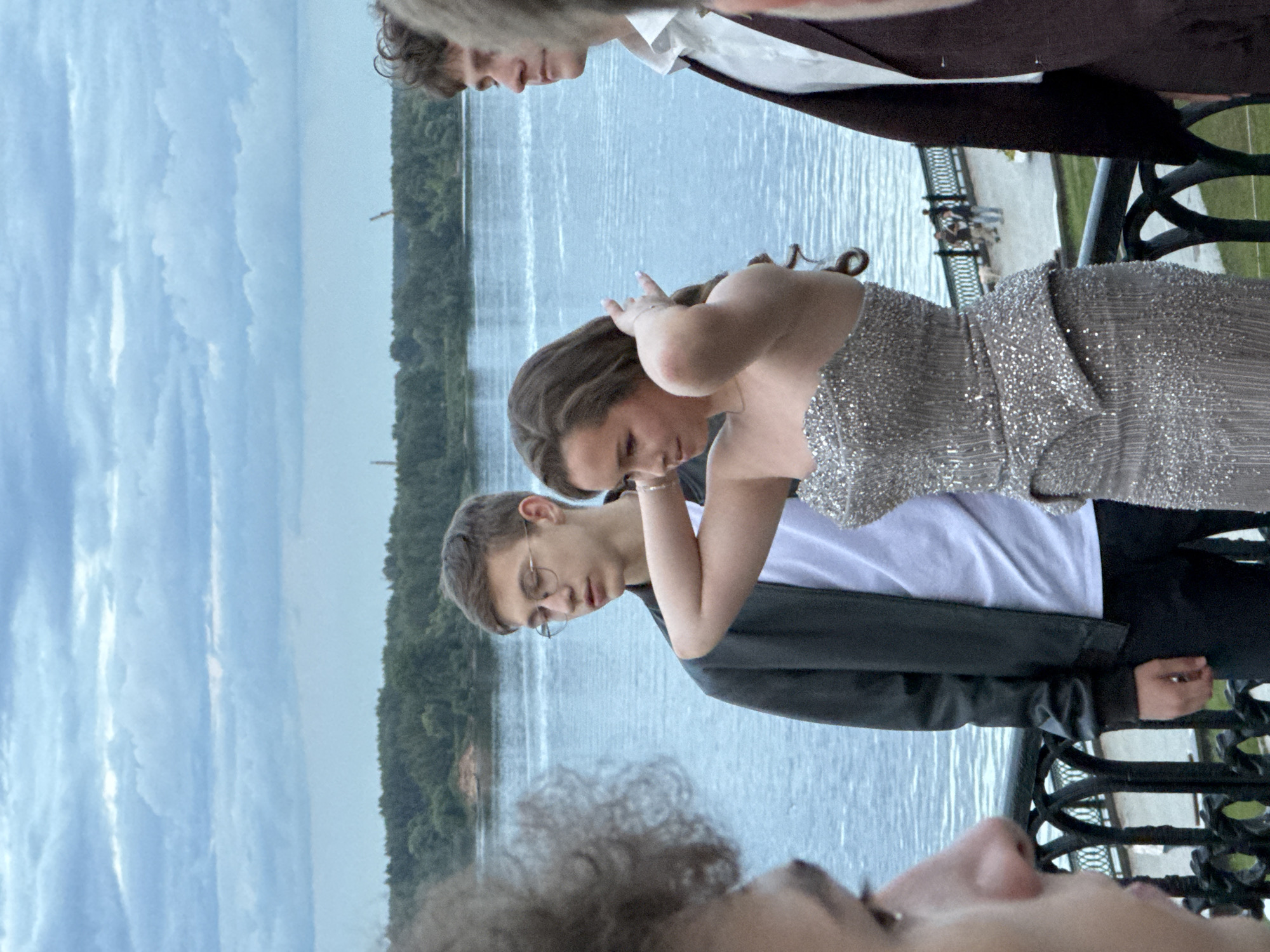 This media file depicts a group of young people on a balcony overlooking a body of water. The individuals appear to be dressed for a formal occasion, with one person wearing a sparkly silver dress and another a suit jacket over a white shirt. The scene is set during the daytime, with a cloudy sky. In the background, a tree-lined shore is visible, suggesting a natural or park-like setting. The people are interacting with each other, with one person adjusting their hair and another looking down, possibly in conversation. The overall mood appears to be relaxed and social. The location is identified as Шебунино, Russia. There is no visible text in the media file.