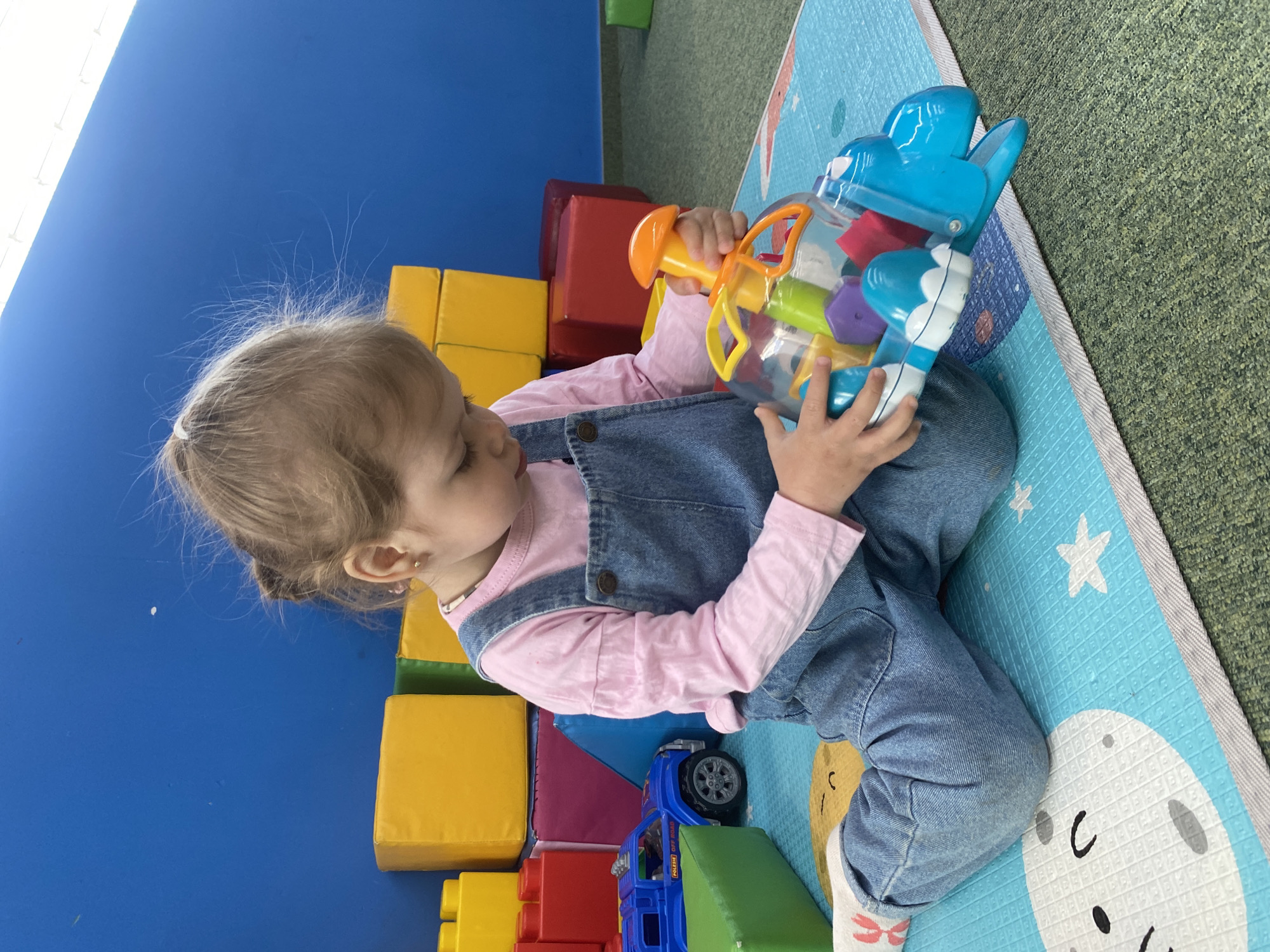 A young child with blonde hair pulled back in a ponytail is sitting cross-legged on a blue foam play mat with moon and star designs. The child is wearing denim overalls over a pink long-sleeved shirt, and socks with pink bows are visible on the feet. The child is holding a clear plastic toy filled with colorful geometric shapes, the toy also has blue bunny ears on the top. To the left of the child, there is a stack of foam blocks in yellow, red, and purple. A blue toy truck is visible near the blocks. The background includes a bright blue wall and green carpet. The city of Perm, Russia cannot be confirmed.