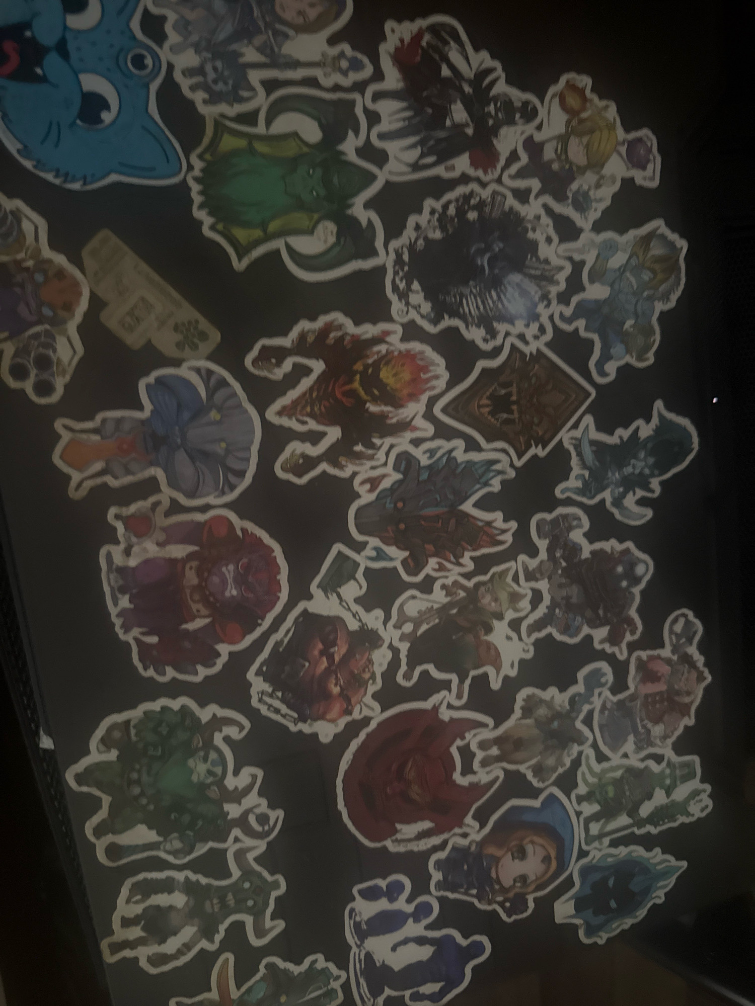 The image is a close-up, dimly lit, overhead shot of a collection of stickers adhered to a dark surface, possibly a notebook or a wall. The stickers depict various cartoon-style characters, many of which appear to be from fantasy or gaming genres. There are characters with dragon-like features, armored figures, and stylized humanoids. The colors are vibrant in some stickers, while others are muted due to the lighting. The stickers are arranged in a somewhat haphazard manner, with some overlapping. No specific location in Gorzów, Poland is identifiable from the image content.