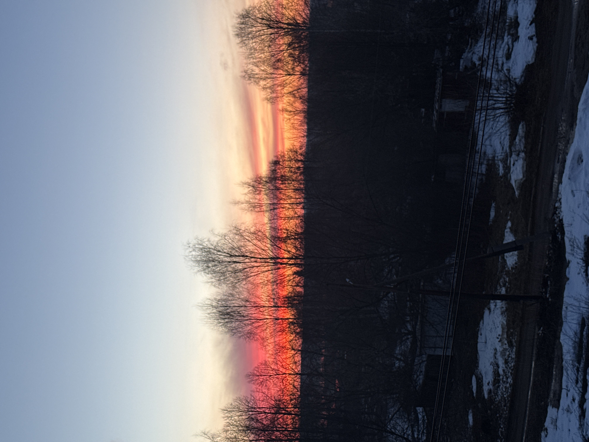 The image depicts a winter sunset in a location that is likely Chaikovsky, Russia. The sky transitions from a pale blue to vibrant hues of orange and red near the horizon. Silhouetted trees form a dark band across the middle of the frame. The foreground on the right side shows a snow-covered landscape, along with some power lines and utility poles. A dark road or path is visible alongside the snow. No people are visible. The environment is cold and characteristic of a late winter or early spring scene.