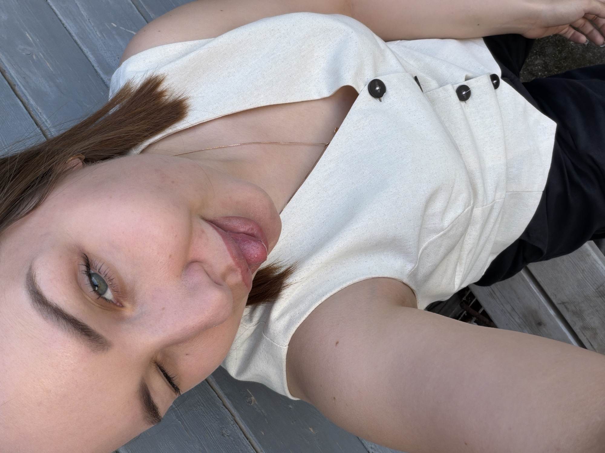 A person is lying on a wooden surface. The person has light skin, brown hair, and green eyes. The person is wearing a light-colored, button-down top and black pants. The person's tongue is sticking out slightly. The wooden surface appears to be part of a deck or platform. No information is available that identifies the location as Chaikovsky, Russia.