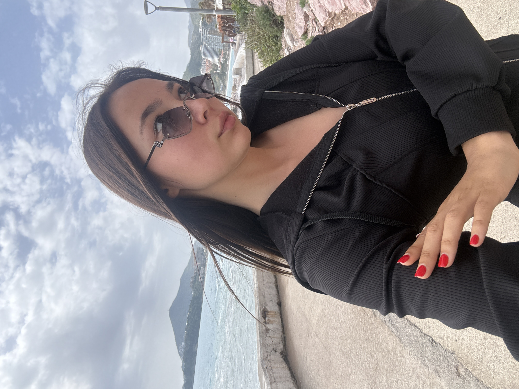 A woman poses outdoors on a concrete path adjacent to a body of water and a cityscape. The woman, wearing a black zip-up hooded jacket and sunglasses, looks away from the camera. She has straight, dark hair and wears red nail polish. The cityscape includes buildings and vegetation on elevated ground. The sky is overcast. The location is in the city of Unknown, Unknown.