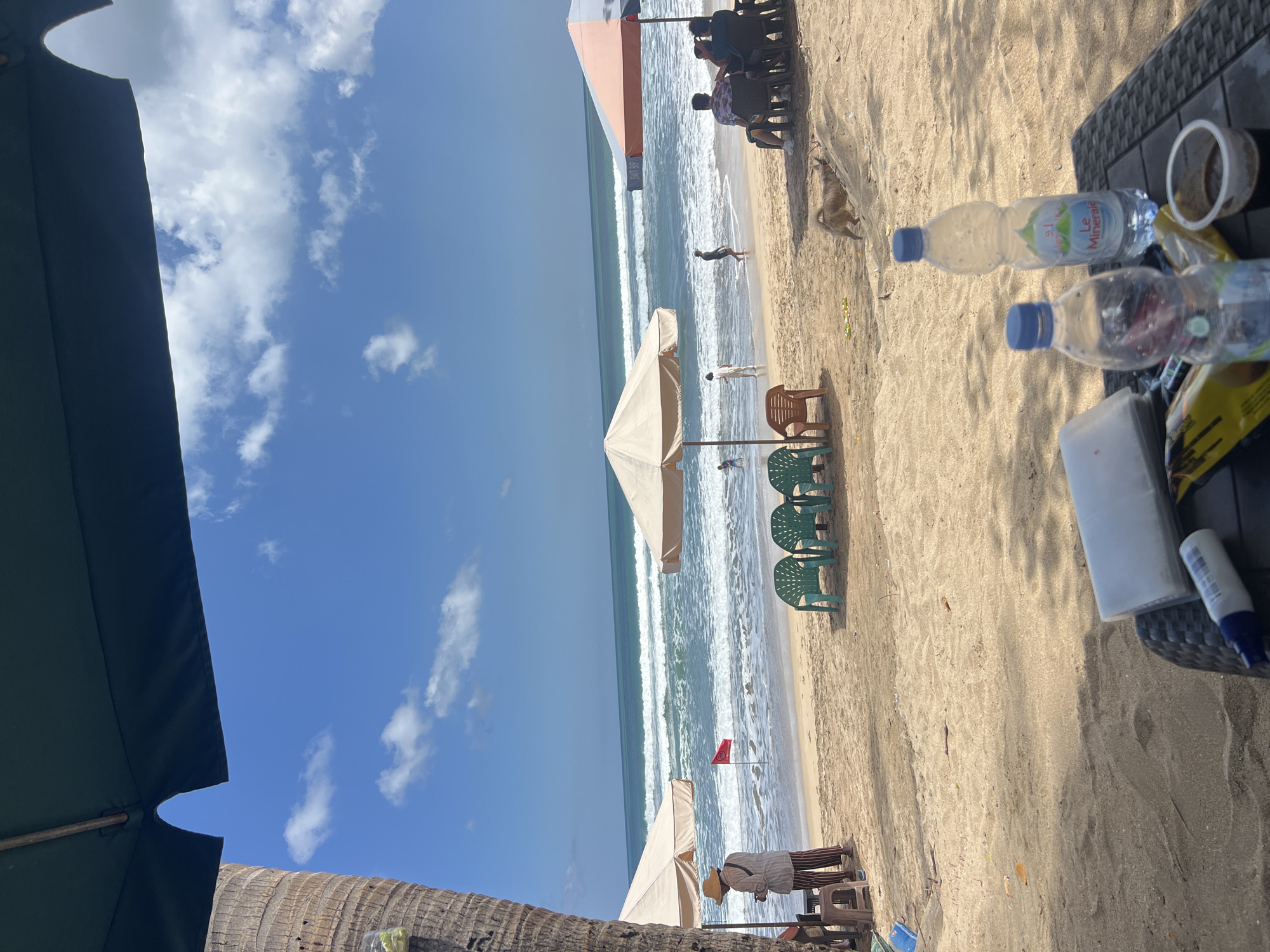 The image depicts a beach scene in Kuta, Indonesia, on a sunny day with blue skies and scattered clouds. In the foreground, a table is set up with two plastic water bottles, a small container of coffee, and a packet of tissues. Beyond the table, a sandy beach stretches out towards the ocean, with gentle waves rolling onto the shore. Several beach umbrellas are set up, providing shade, and a few rows of green plastic chairs are neatly arranged on the sand.
In the middle ground, there are a few people on the beach. One person in a hat and striped pants stands near an umbrella and a small table, appearing to interact with someone or something out of frame. A red flag is planted in the sand near the water's edge, indicating a swimming area. Further out in the water, a few individuals are visible, some appearing to be swimming or wading.
In the background, the ocean meets the horizon under the bright sky. On the right side of the image, another umbrella is visible, under which a group of people are seated at a table. A small dog is seen in the sand near the foreground, walking away from the camera. The overall atmosphere suggests a relaxed beach environment, likely a popular tourist spot in Kuta.