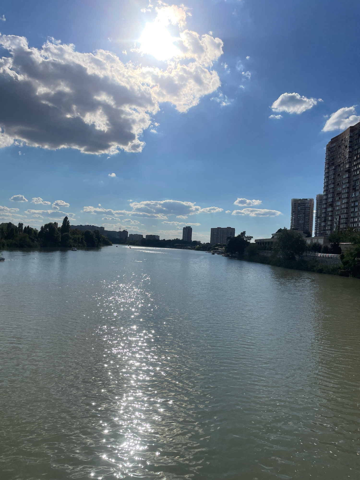 The image depicts a wide river with a cityscape visible on both banks under a bright, sunlit sky with scattered clouds. The sun is high in the sky, casting a strong glare on the water's surface, creating a shimmering effect with numerous bright reflections. On the right bank, several tall, modern apartment buildings with many windows dominate the skyline. On the left bank, there is a line of trees, with some smaller buildings interspersed further back. A few boats are visible on the water, and the overall atmosphere is serene and summery. This scene is located on the Kuban River in the city of Krasnodar, Russia, likely near the city center.