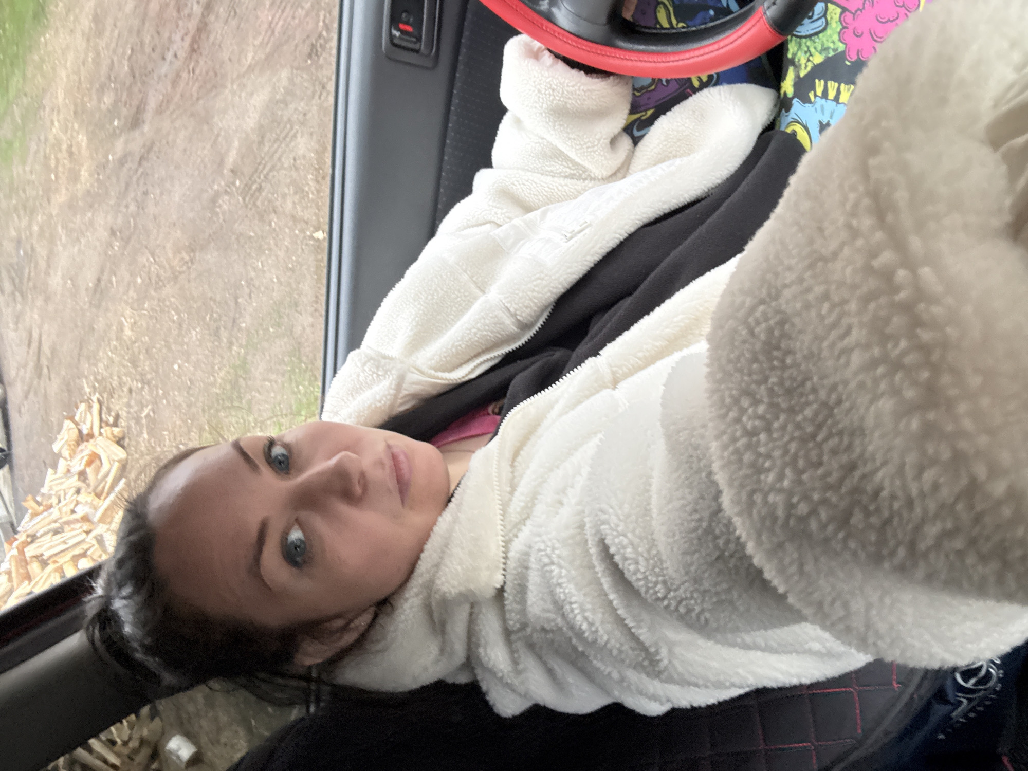The image, rotated 90 degrees counter-clockwise, depicts a woman taking a selfie from inside a vehicle. The woman has dark hair pulled back, prominent blue eyes, and a neutral expression, looking directly at the camera. She is dressed in a white, fluffy zippered jacket over what appears to be dark clothing, with a glimpse of a pink garment underneath.
The setting is the interior of a vehicle, indicated by a red steering wheel cover, a dark car door panel with a control switch, and a black quilted seat or surface with red stitching. Behind the steering wheel, a colorful fabric with abstract, cartoon-like patterns in blue, yellow, black, and pink is visible.
Through the vehicle window on the left, an outdoor scene is visible. The ground appears to be a dry dirt or gravel path, showing faint tire marks and sparse green patches. Further to the left and slightly below, there is a pile of what looks like cut wood or kindling. The natural light suggests it is daytime. The scene outside implies a rural or semi-rural environment, consistent with the provided location context of Chaikovsky, Russia. There is no visible text in the image.