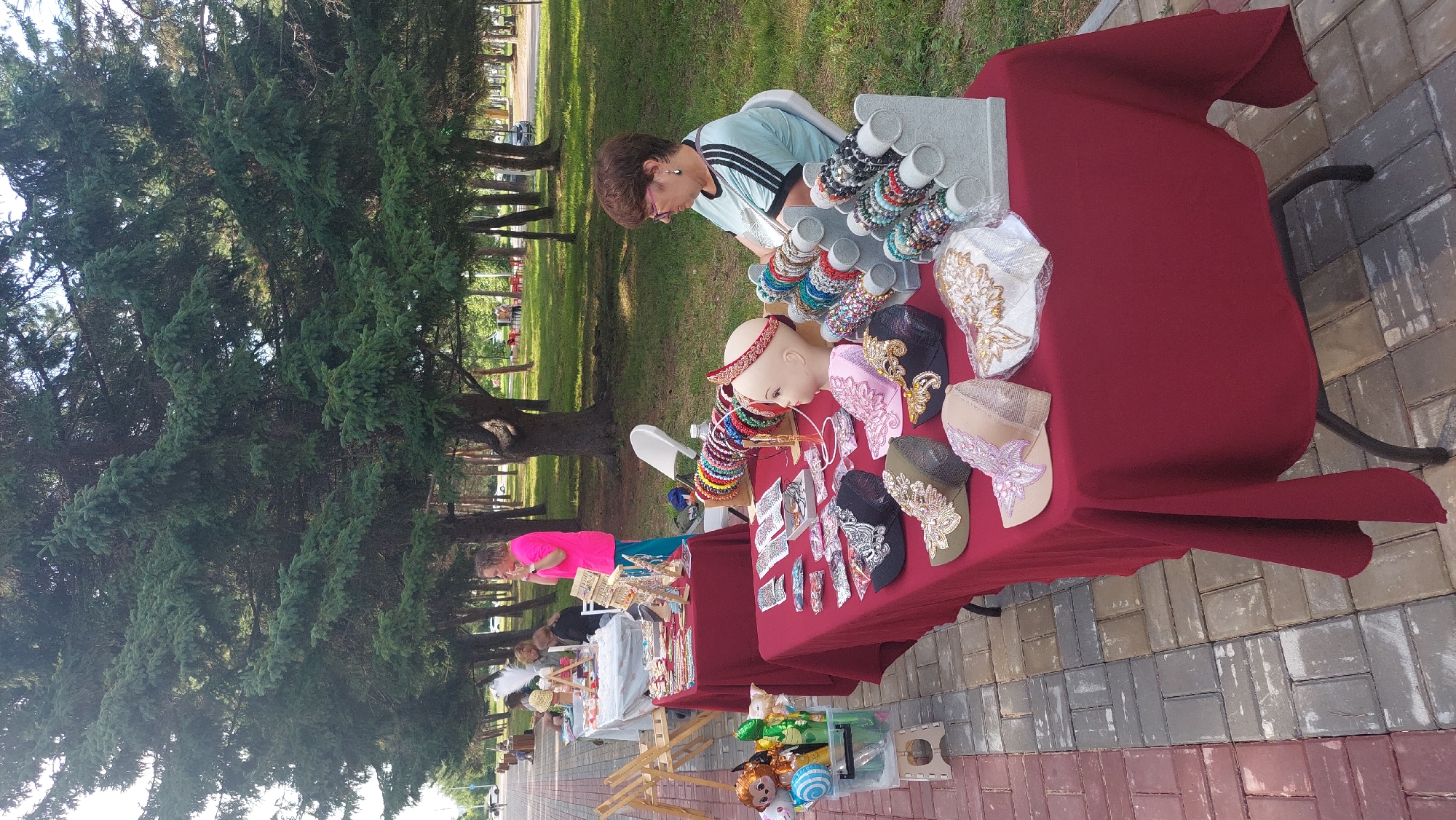 This image captures an outdoor craft fair scene, likely in a park in Komsomolsk-on-Amur, Russia. In the foreground, a table draped with a burgundy cloth displays an assortment of handmade accessories. These include decorative caps with intricate embroidery and embellishments, headbands adorned with colorful beads and jewels, and a mannequin head showcasing a headband. To the left, a display box holds balloons and toys, suggesting items for children.
Further back, a woman with short brown hair and glasses is seated at another table, which is also laden with similar handmade jewelry and accessories, primarily bracelets displayed on stands. She appears to be attending to the stall. Behind her, another woman in a bright pink top and teal pants stands near a table displaying more crafts, actively engaged in conversation or making a sale.
The setting is characterized by lush green trees with dense foliage providing shade, and a paved walkway made of interlocked pavers in shades of red, grey, and beige. The overall atmosphere suggests a sunny day, with people browsing and purchasing unique, handcrafted items. The presence of other tables and people in the background indicates a larger market event. The environment suggests a public park or recreational area commonly found in Russian cities.