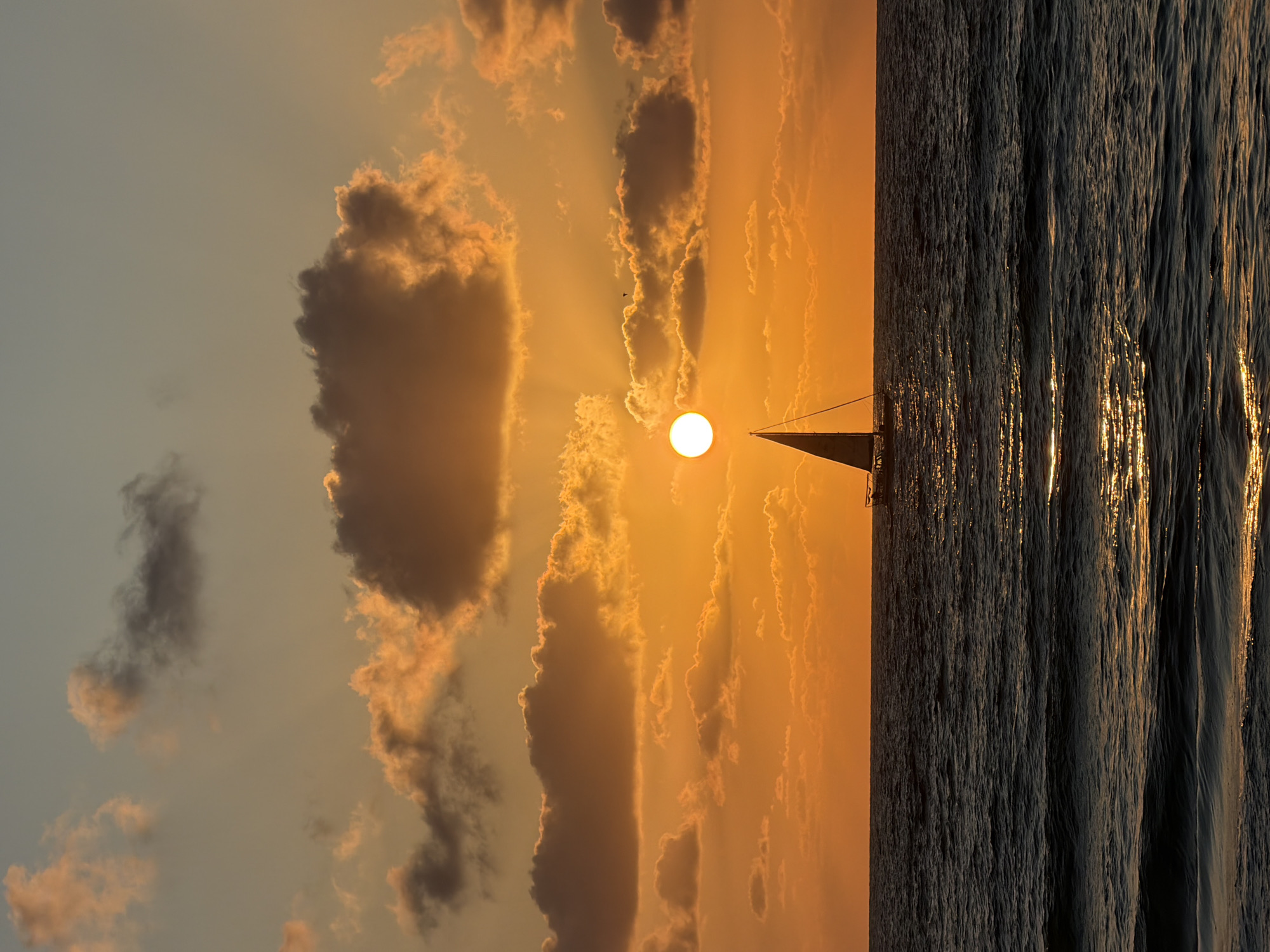 The image captures a vertical shot of a sunset over the Mediterranean Sea, with a sailboat silhouetted against the setting sun, likely off the coast of Tel-Aviv, Israel. The sky is a blend of orange and yellow hues, with scattered clouds partially obscuring the sun, creating beams of light. The sea appears dark and textured, with reflections of the sunlight shimmering on the water's surface. The sailboat's mast and sail are visible as a dark outline against the bright sun. There are no people visible in the image.