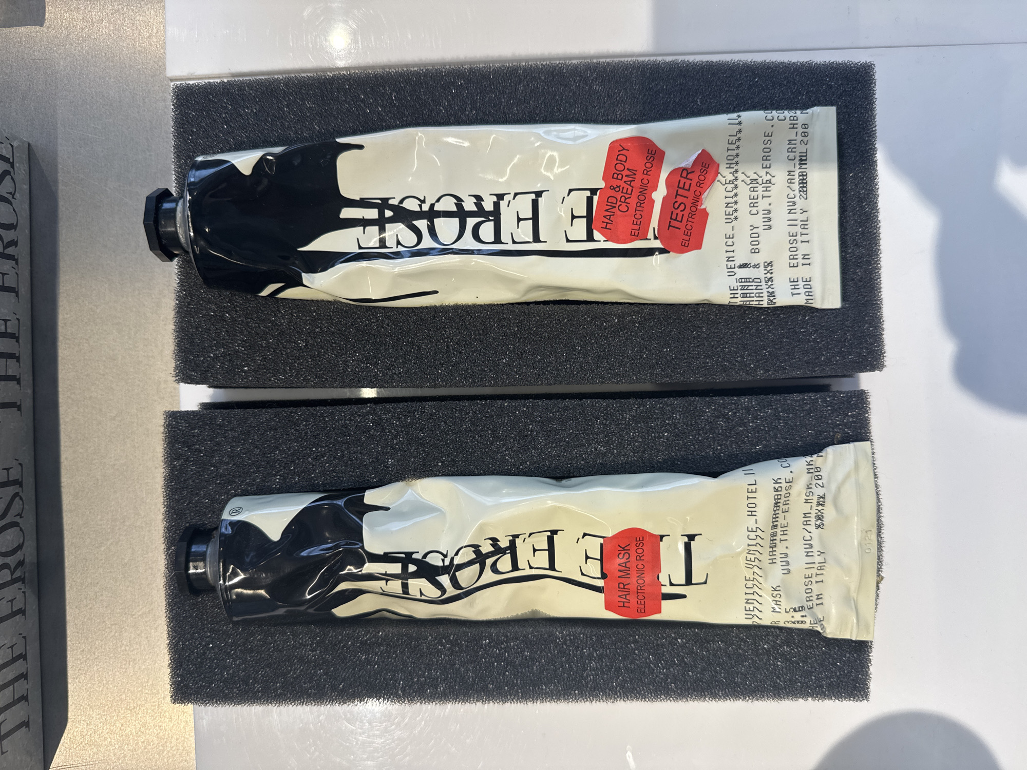 The image shows two tubes of beauty products displayed in what appears to be a retail setting. Both tubes are white with black ink blot style patterns. The top tube is labeled "THE EROSE HAND & BODY CREAM ELECTRONIC ROSE" and has a red tag indicating it is a "TESTER." The bottom tube is labeled "THE EROSE HAIR MASK ELECTRONIC ROSE." The tubes rest in black foam inserts. The label includes "VENICE-VENICE-HOTEL IL" and "MADE IN ITALY." Based on the labels, this would likely be available at The Venice Venice Hotel in Italy. The city of Milan, Italy, is not specifically mentioned in the image but is simply implied because it's made in Italy.