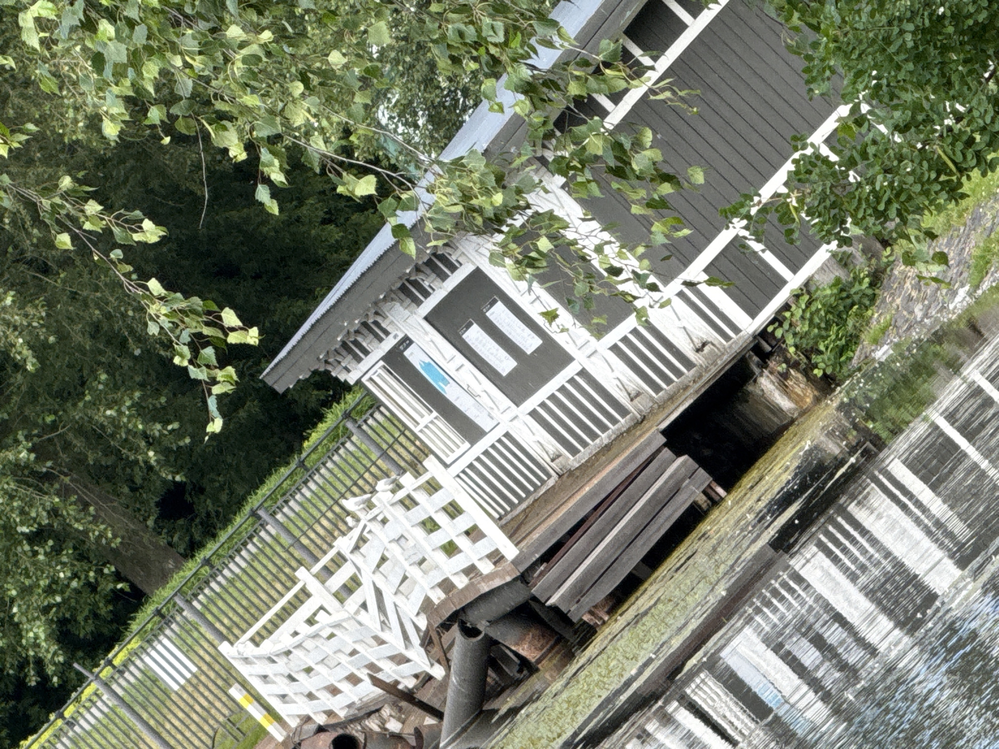 The image shows a building with a dark gray exterior and a white lattice fence. The building has a metal roof and several windows. The fence surrounds a wooden platform that extends over the water. There are trees and foliage in the background. The image is taken at an angle, giving the impression of a tilted perspective. There are no people or animals visible in the image.