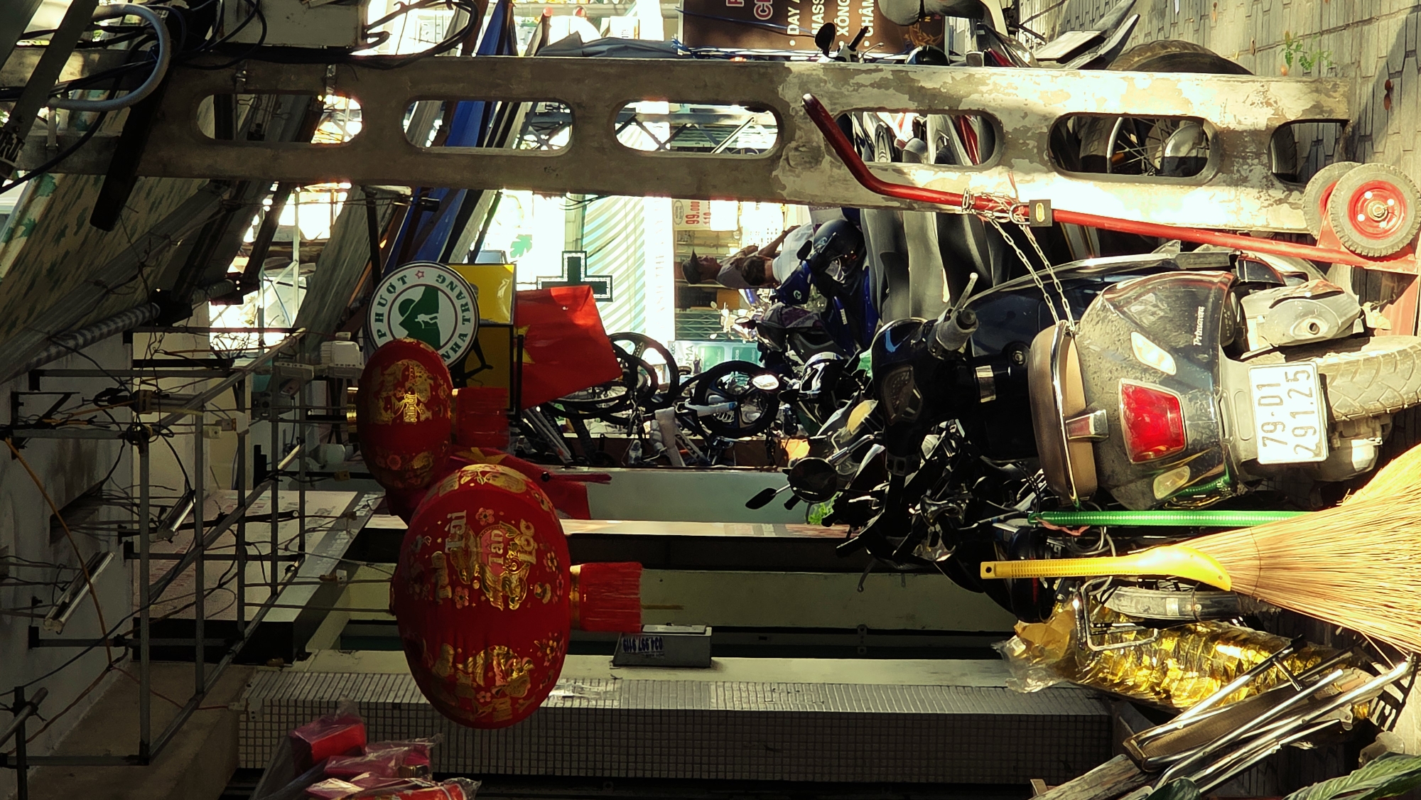 This eye-level shot, taken in Nha Trang Ward, Vietnam, captures a chaotic scene of stacked objects. Dominating the right side is a grey Primavera scooter, oriented horizontally with its tail light and license plate (79-01 291.25) visible. Nearby, a tan broom leans against a stack of what appears to be gold-wrapped items, possibly merchandise. Above the scooter is a metal structure with cutout rectangles and red wheels.
The left side features a series of metal shelves with red boxes. Ornate red lanterns hang from the shelves. A round sign with Vietnamese script, including "PHƯƠT" and "NHA TRANG", is visible. The background reveals more parked scooters, red flags, and various merchandise. In the distance is a sign advertising massage services and foot care, as well as a price sticker of 99.000 - 110.