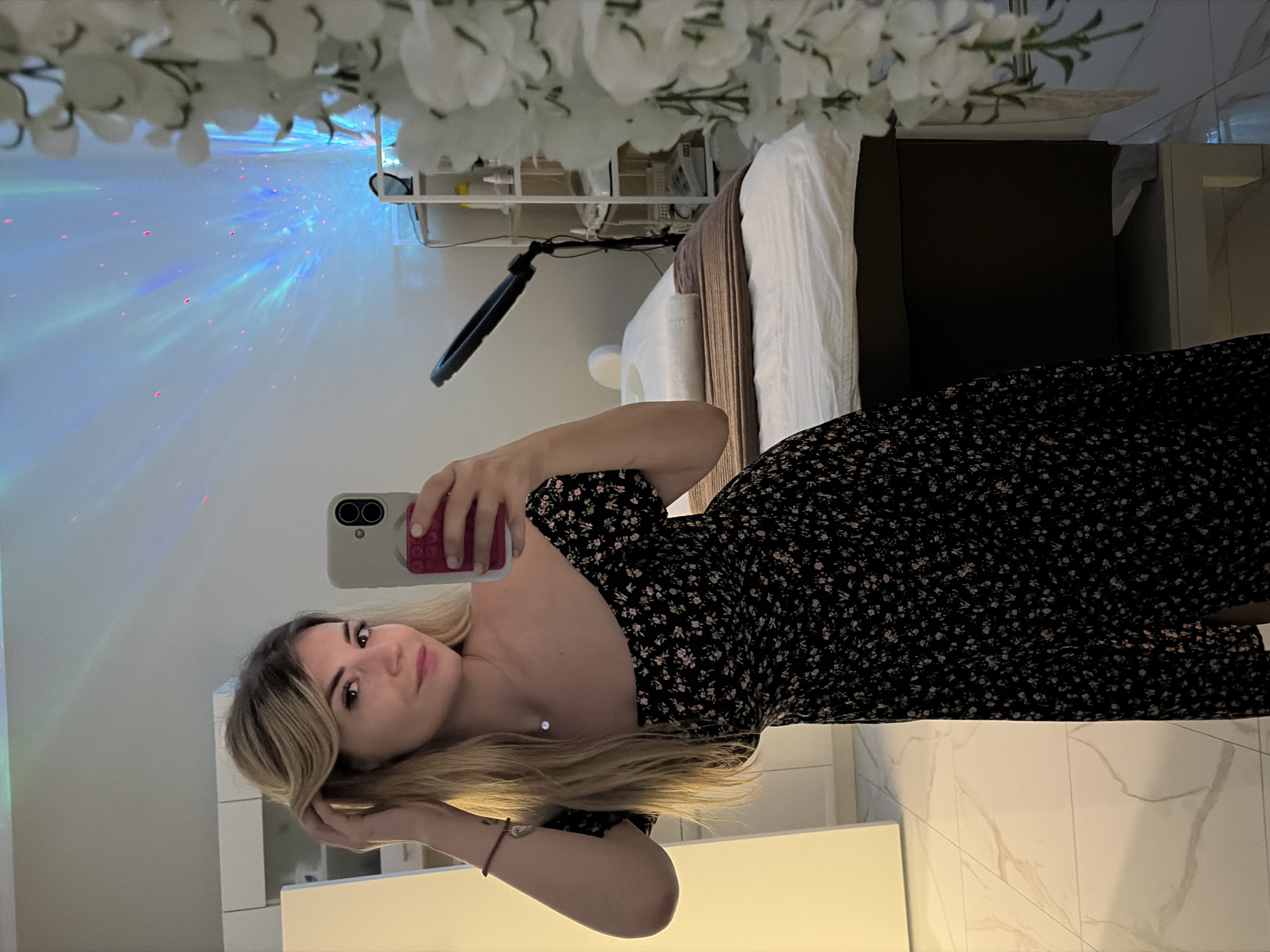 A woman with blonde hair, wearing a black floral off-the-shoulder dress, takes a selfie in a room that appears to be a spa or treatment room. She is holding a phone with a pink pop socket. The room contains a massage table with white linens and a brown throw blanket, a shelf unit with various bottles and equipment, and a ring light. The wall behind her is illuminated with a colorful star projector. The floor is tiled with a marble pattern. The overall atmosphere is calm and relaxing. This scene is located in Budva, Montenegro.