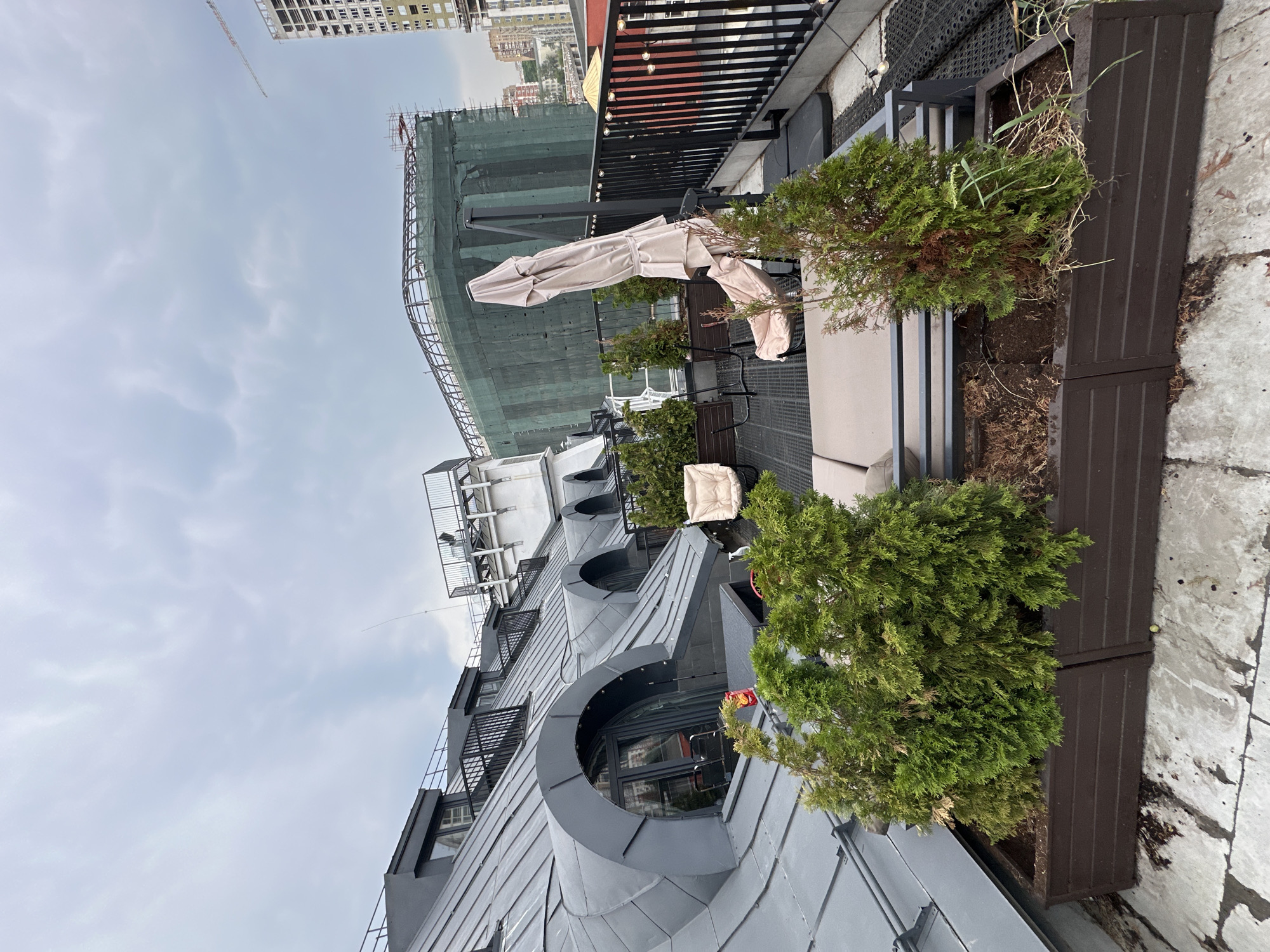 This is a medium-shot, eye-level photograph of a rooftop terrace in Moscow, Russia, under a cloudy sky. The terrace features dark gray metal roofing with arched dormer windows, a black metal railing, and dark brown planters filled with green evergreen shrubs. In the foreground, a beige lounge chair and a beige umbrella are visible. Beyond the terrace, there is a large building under construction, covered in green netting. To the right, a tall, modern building is partially visible. The overall impression is one of an urban outdoor space, likely part of a residential or commercial building, with ongoing construction in the background.